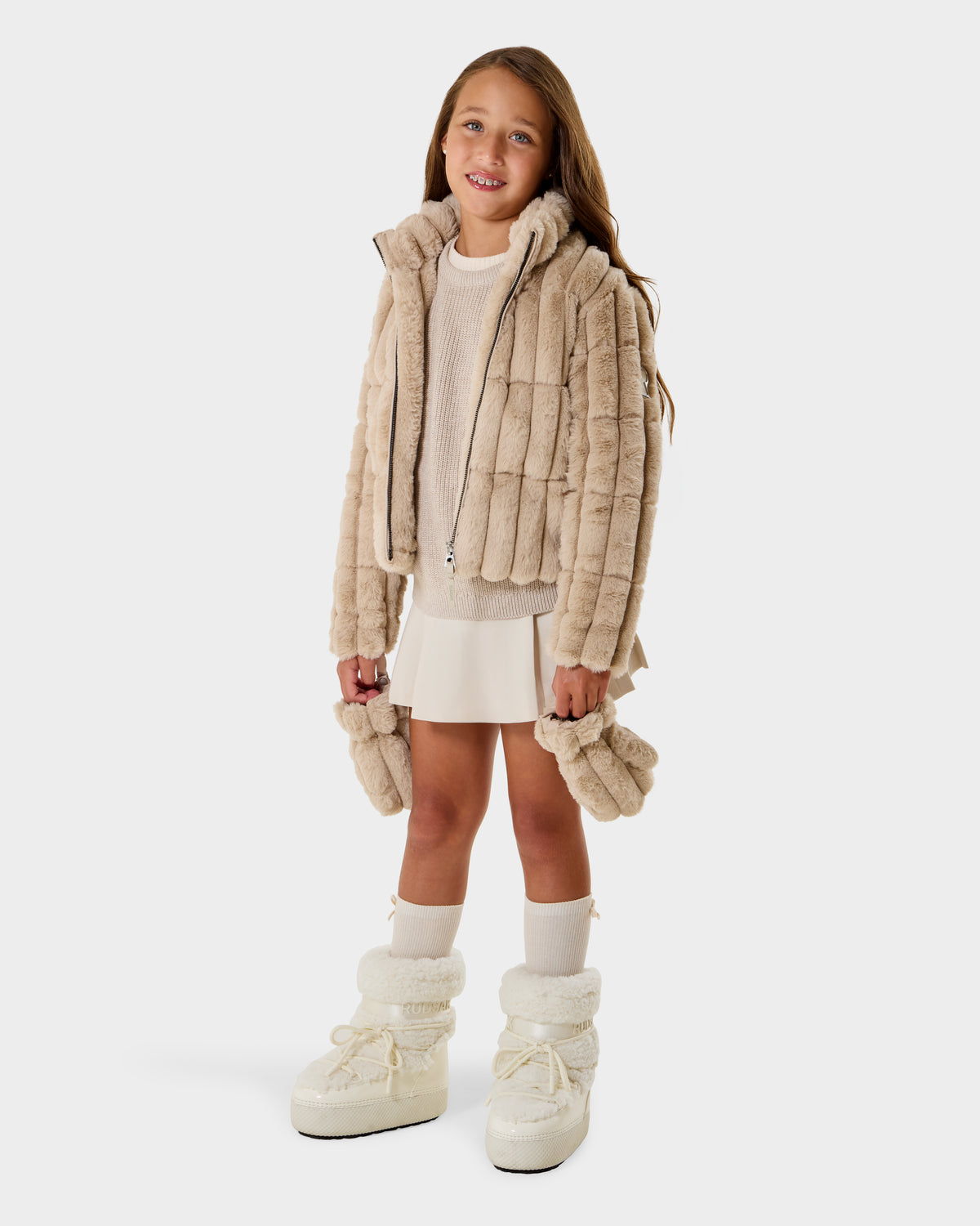 A young girl wearing the Rudsak ANASTASIA K - 0125000 Vanilla jacket smiles against a plain white background, styled with matching mittens, a cream sweater, a white skirt, and winter boots.