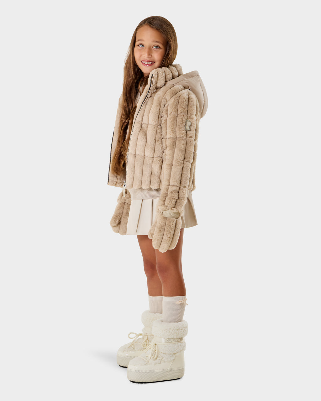 A young girl with long brown hair wears the Rudsak ANASTASIA K - 0125000 Vanilla faux fur shell jacket, white pleated skirt, knee-high socks, mittens, and white boots, standing and smiling against a plain white background.