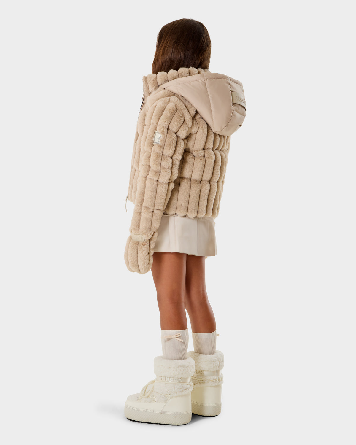 A young girl with long brown hair, seen from behind, wears the Rudsak ANASTASIA K - 0125000 Vanilla puffer jacket with down insulation, a cream skirt, beige mittens, white socks, and off-white boots against a plain background.