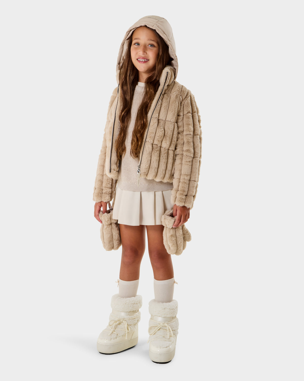 A young girl with long brown hair wears the Rudsak ANASTASIA K - 0125000 Vanilla beige faux fur shell jacket with a hood, layered over a cream sweater and white pleated skirt, paired with white socks and boots against a plain white background.