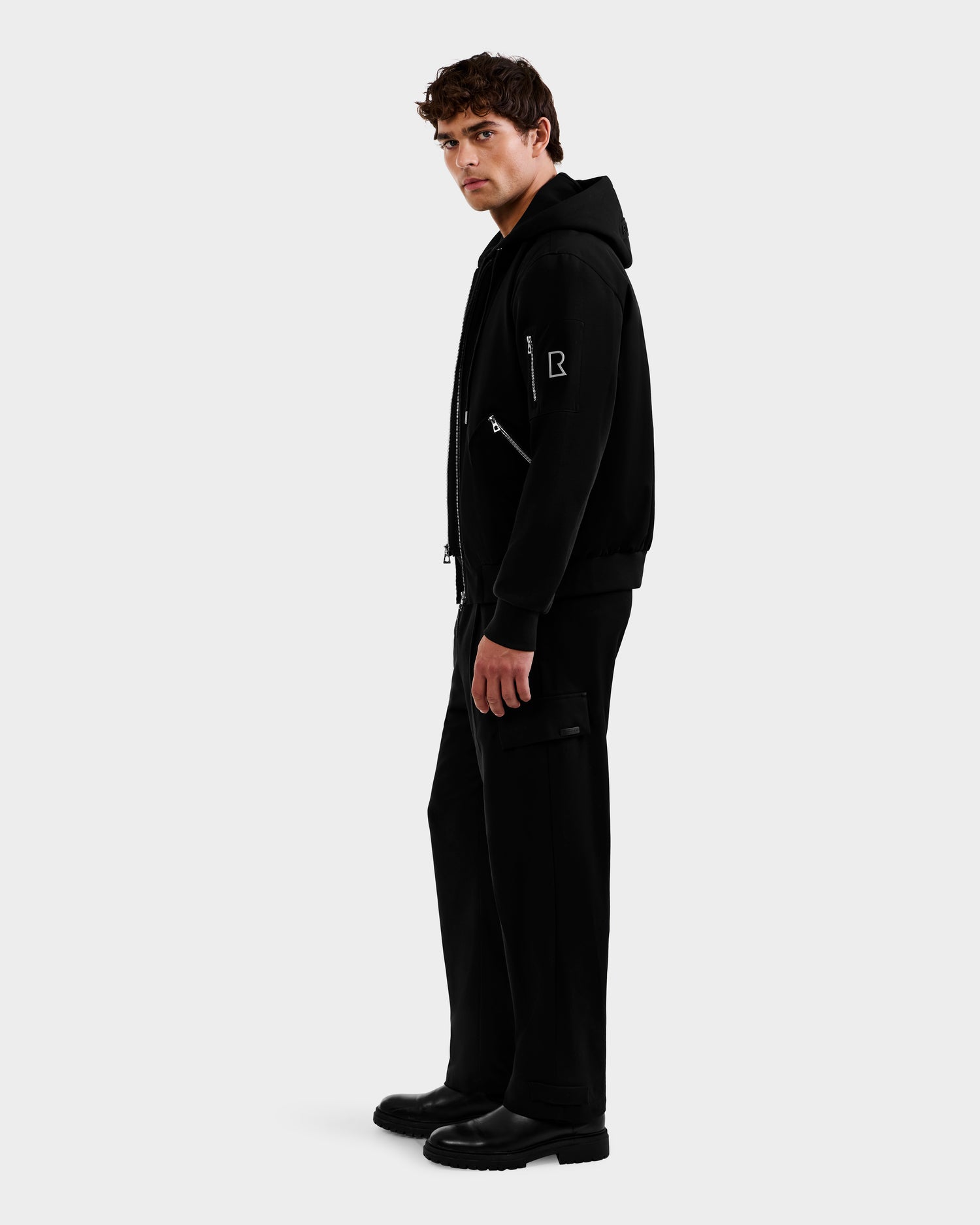 A man wearing the Rudsak ANDRE - 6124020 Black zip-up jacket with removable hooded bib, black pants with side pockets, and black boots stands against a plain white background, glancing over his shoulder.