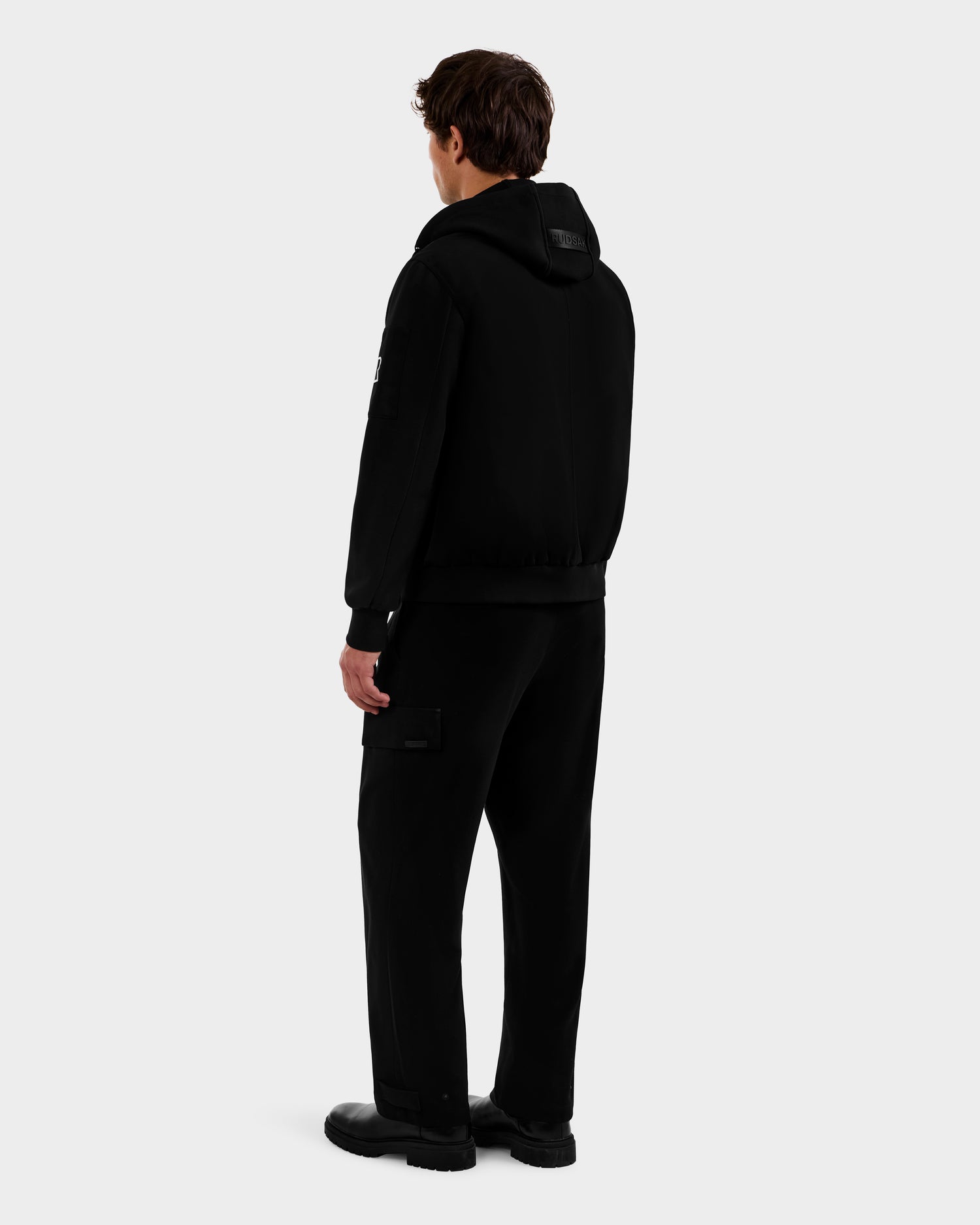 A person stands facing away, wearing the Rudsak ANDRE - 6124020 Black men's lightweight bomber jacket with a removable hooded bib, paired with black side-pocket pants and black shoes, against a plain light gray background.