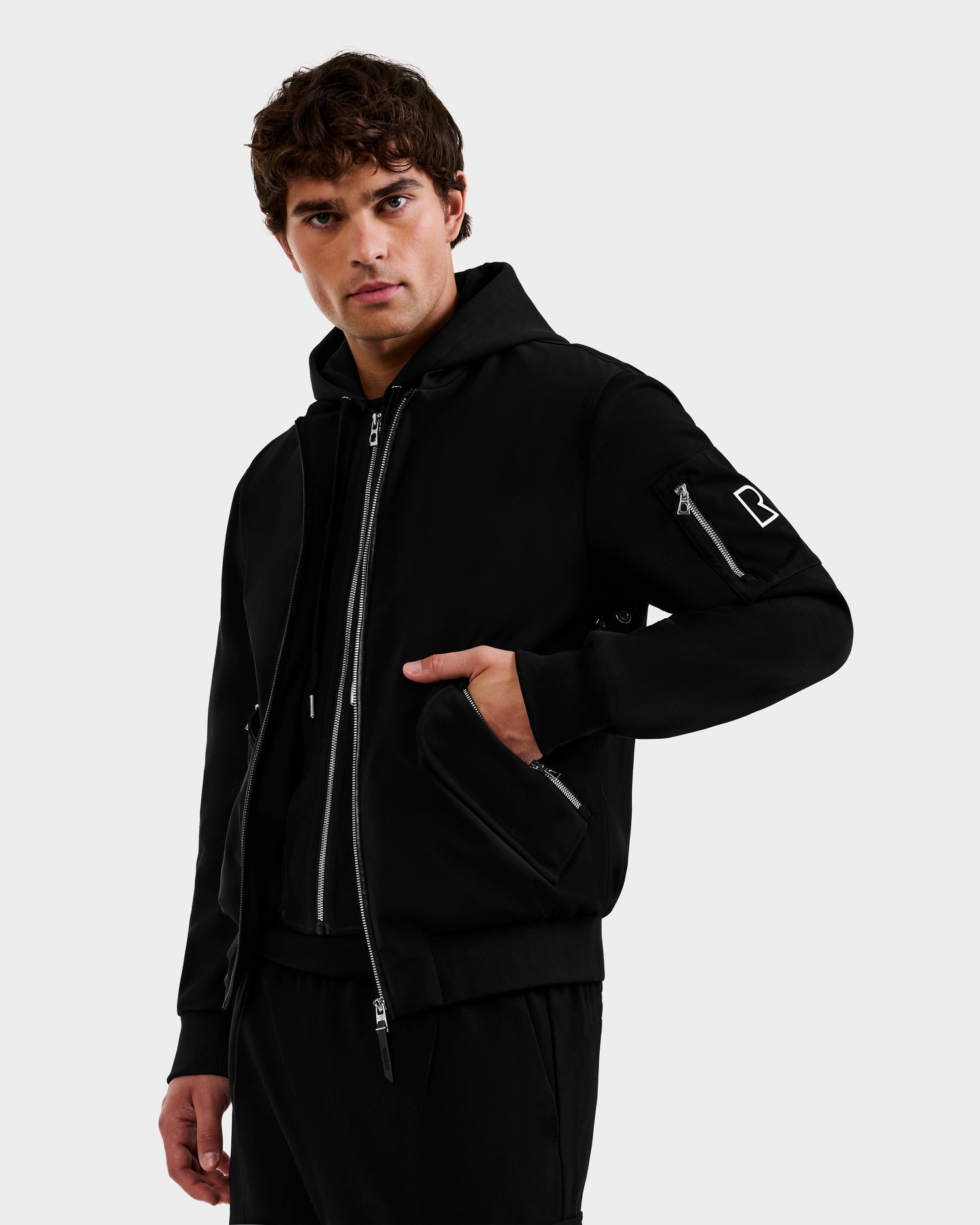 A young man with wavy brown hair wears the Rudsak ANDRE - 6124020 Black men's lightweight bomber jacket, featuring a removable hooded bib, with one hand in his pocket against a plain light gray background.