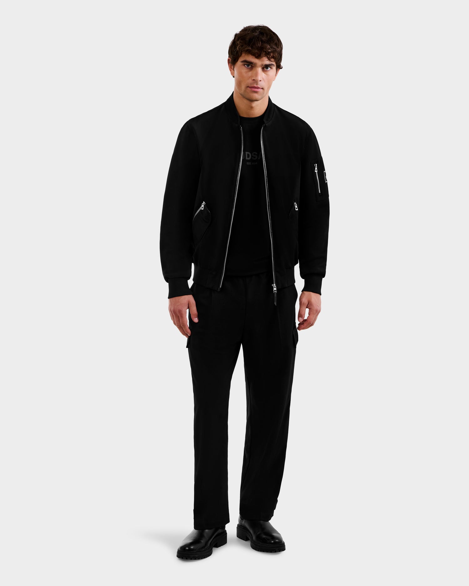 A man stands against a light background, wearing the Rudsak ANDRE - 6124020 Black men's lightweight bomber jacket with removable hooded bib, paired with a black T-shirt, pants, and shoes. He faces forward neutrally with his hands at his sides.