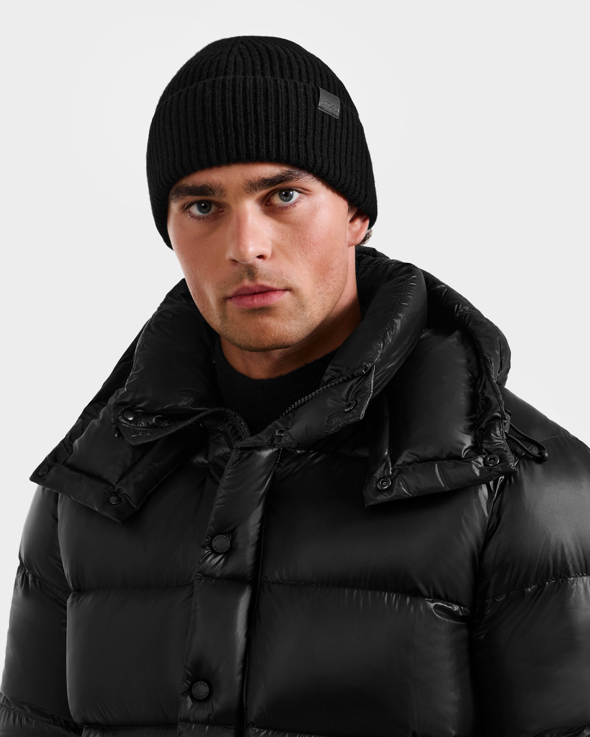 A man wearing the Rudsak ANDREY - U425507 Black puffer jacket and a black rib-knit beanie, looking directly at the camera against a plain light background.