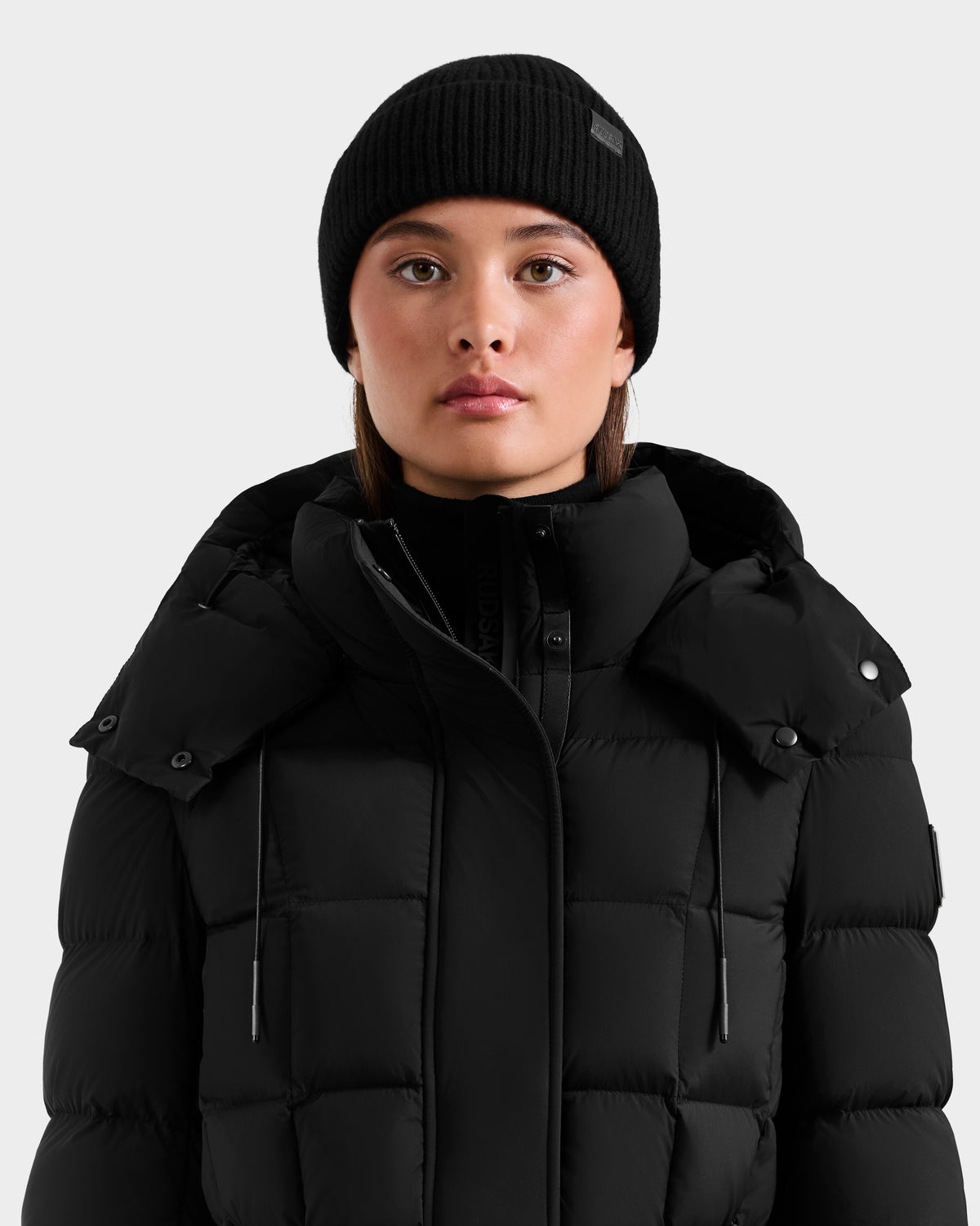A person faces forward against a plain light gray background, wearing the Rudsak ANDREY - U425507 Black puffer jacket and a black unisex beanie.