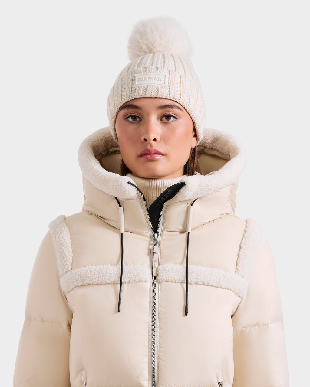 A woman wears the Rudsak ANISA - L423500 Vanilla beige puffer jacket with sherpa-lined hood and a cream knit pom pom hat, standing against a plain background—perfect cozy pieces for any winter wardrobe.