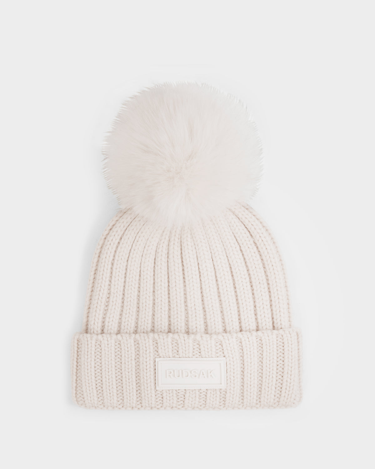 The ANISA - L423500 Vanilla by Rudsak is a cream-colored ribbed knit beanie with a plush faux fur pom pom and folded brim, accented by a rectangular Rudsak label—perfect for elevating your winter wardrobe.