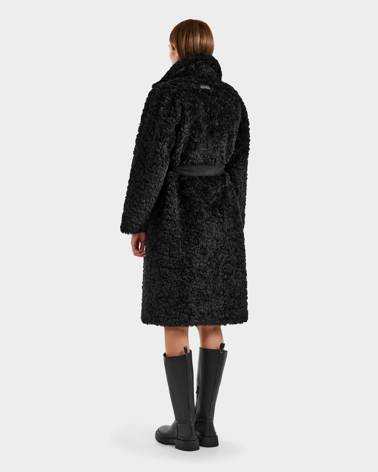 A woman with straight, shoulder-length hair wears the Rudsak ANNABELLE FSU - L125512 Black trench coat and tall black boots, standing with her back to the camera against a plain light background.