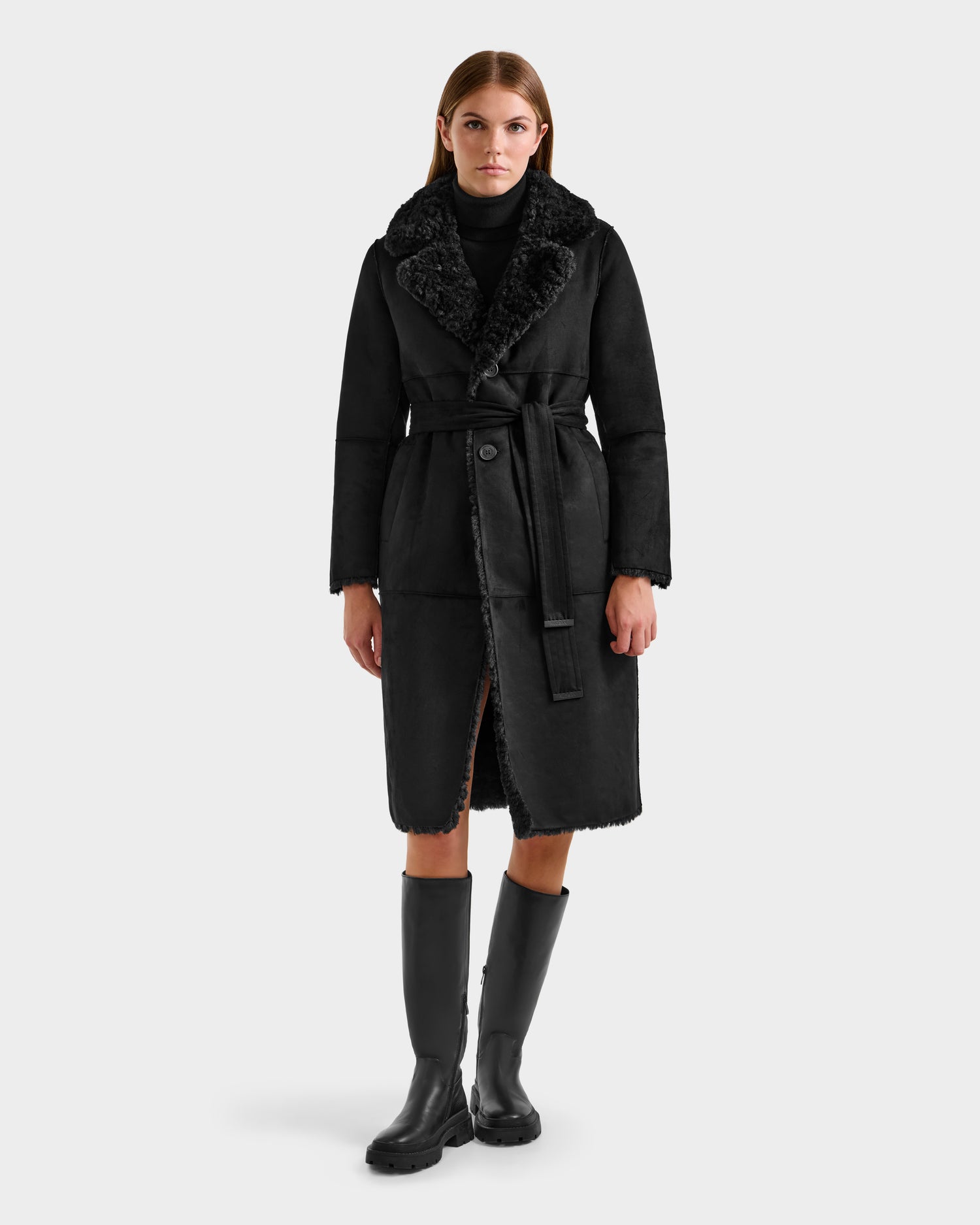 A woman faces forward against a light gray background, wearing the Rudsak ANNABELLE FSU - L125512 Black faux suede trench coat with a tie belt, paired with a black turtleneck and knee-high black boots.