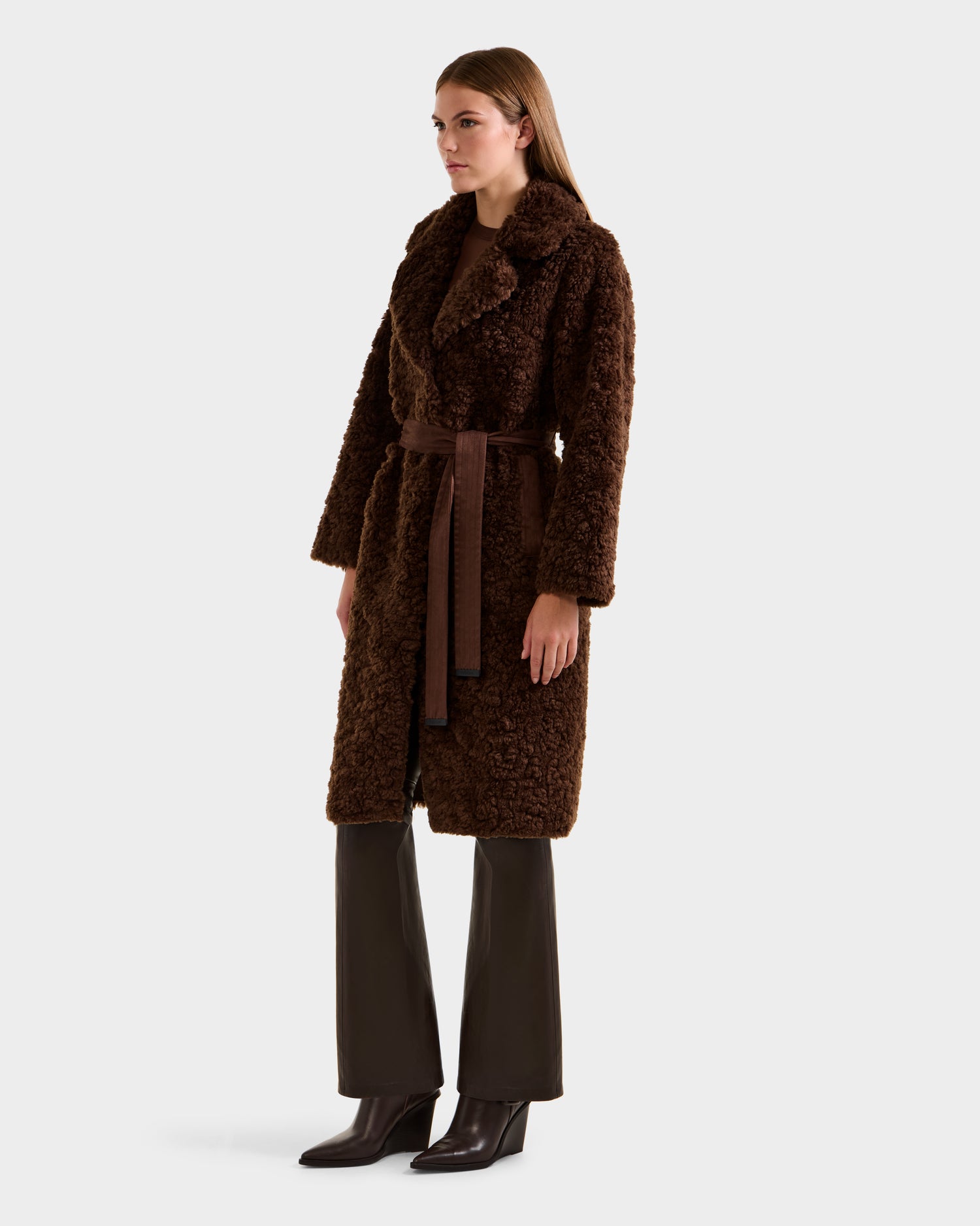A woman stands against a plain light background wearing the Rudsak ANNABELLE FSU - L125512 Espresso faux suede trench coat with a matching belt, styled with brown flared pants and dark heeled boots.