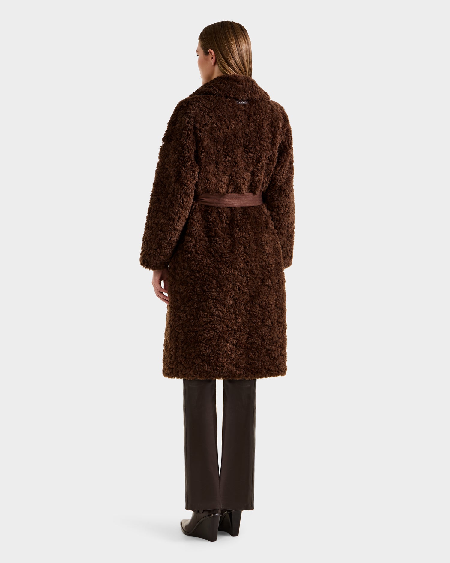 A person with straight brown hair models the Rudsak ANNABELLE FSU - L125512 Espresso sherpa teddy coat, knee-length with a belt, paired with dark pants and black heeled shoes, shown from the back against a plain background.