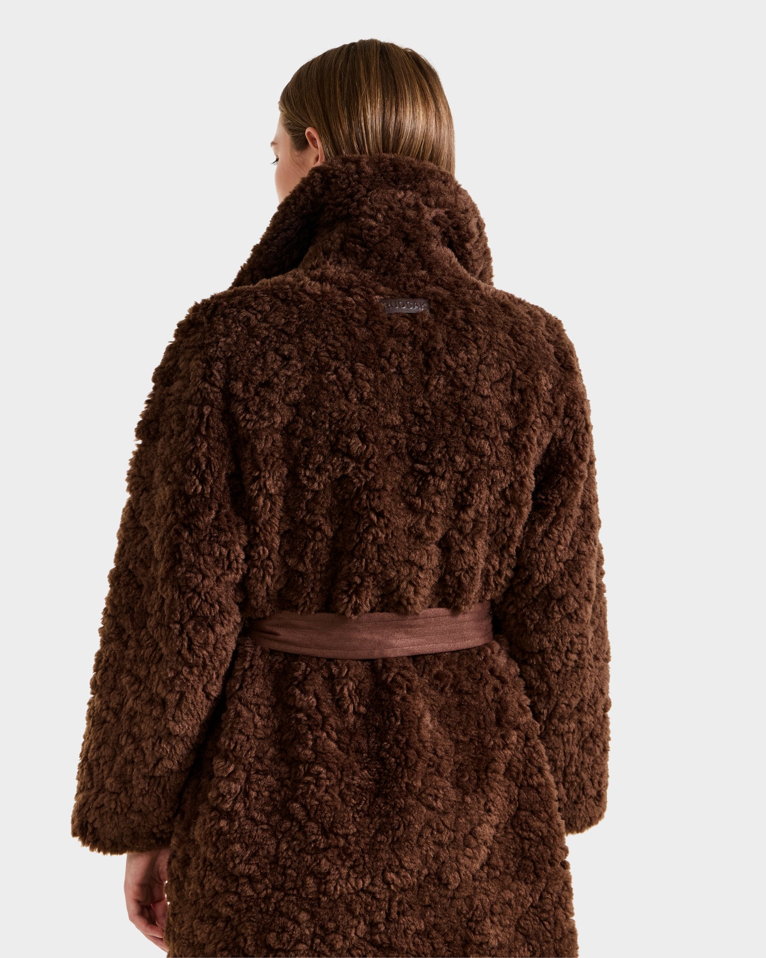 A person with straight, light brown hair is shown from the back, wearing the Rudsak ANNABELLE FSU - L125512 Espresso sherpa teddy coat with a textured finish and matching belt tied at the waist.