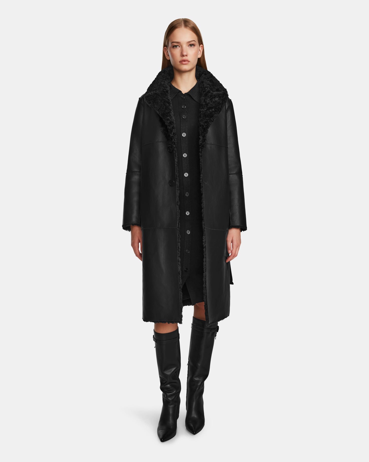 A woman faces forward, wearing the Rudsak ANNABELLE - L124544 Black vegan leather coat over a black dress and knee-high boots, set against a plain white background.