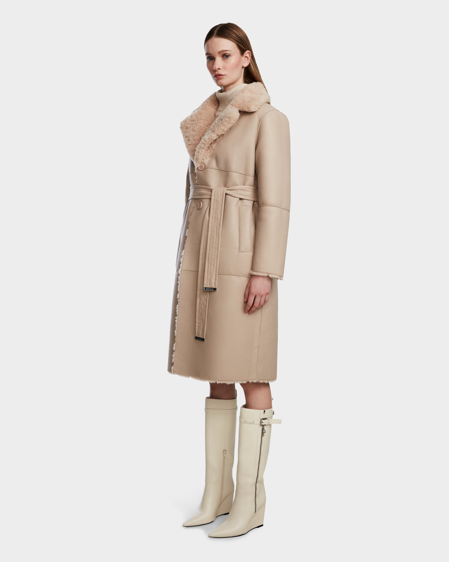 A woman wears the Rudsak ANNABELLE - L124544 Sand long vegan leather coat with a belt and wide collar, paired with knee-high cream boots, standing slightly turned against a plain white background.