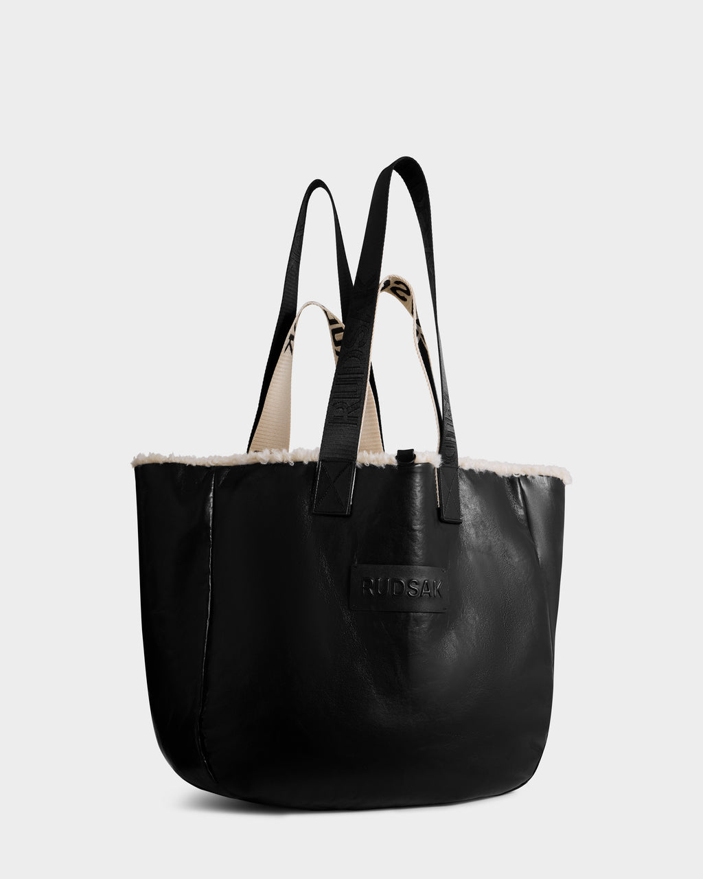 The ANNIAH - 8325541 Black/Vanilla tote bag by Rudsak features a black faux shearling body with a white faux fur trim, two black handles, and an embossed Rudsak logo on the front. Its interior is light-colored and textured.