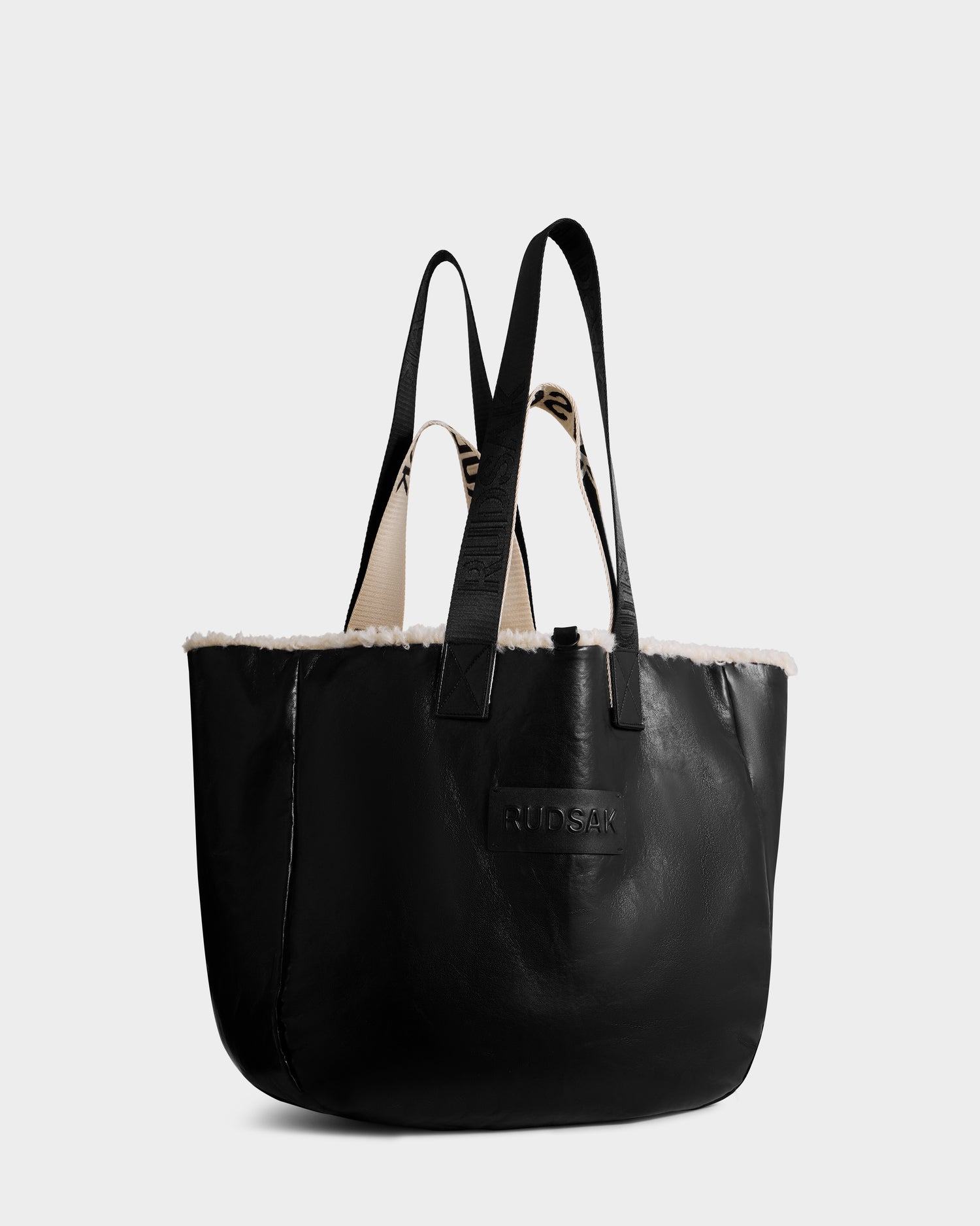 The ANNIAH - 8325541 Black/Vanilla tote bag by Rudsak features a black faux shearling body with a white faux fur trim, two black handles, and an embossed Rudsak logo on the front. Its interior is light-colored and textured.