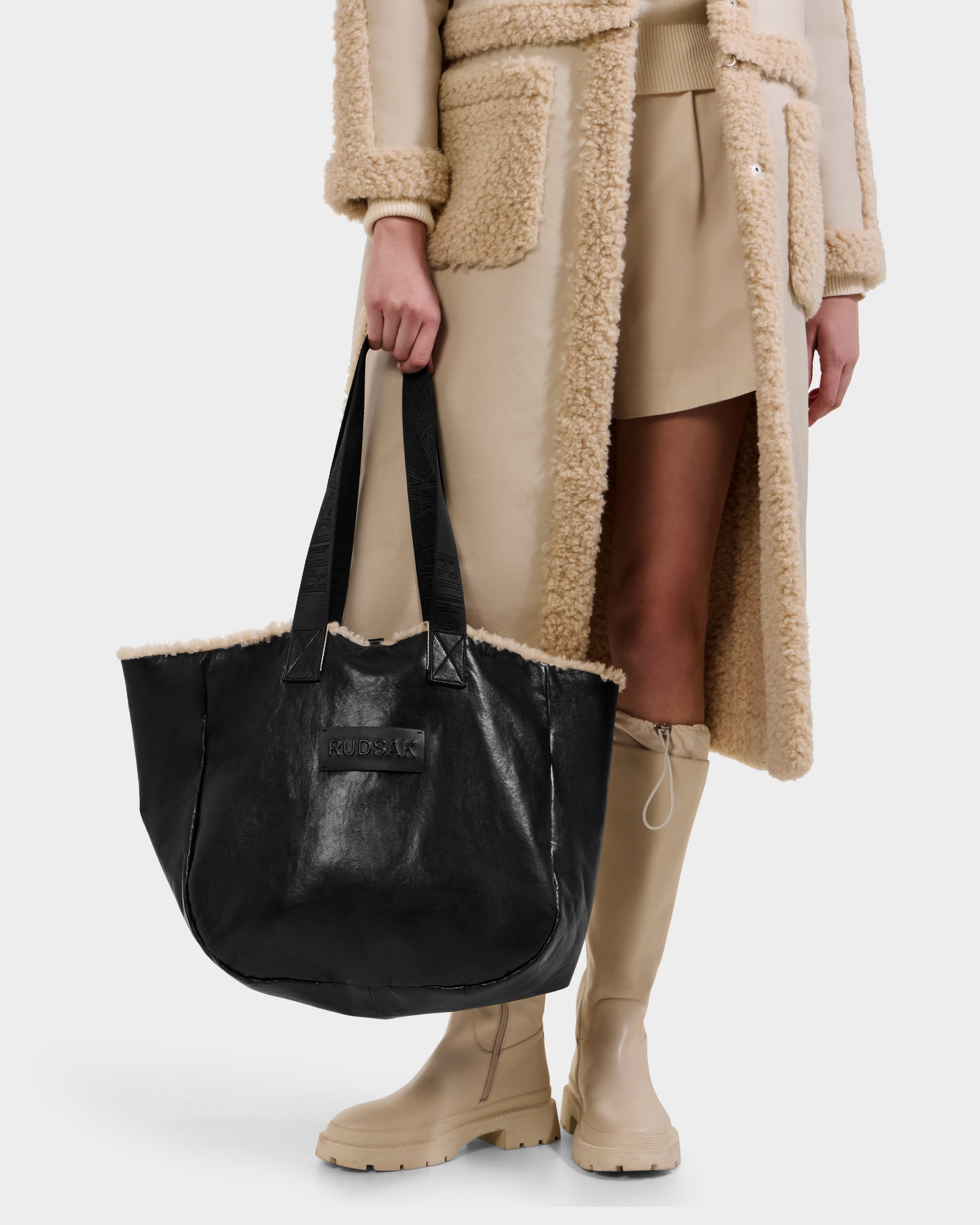 A person wearing a beige shearling coat, skirt, and boots holds the Rudsak ANNIAH - 8325541 tote in Black/Vanilla with plush trim. The photo is cropped to display only the lower half of their body.