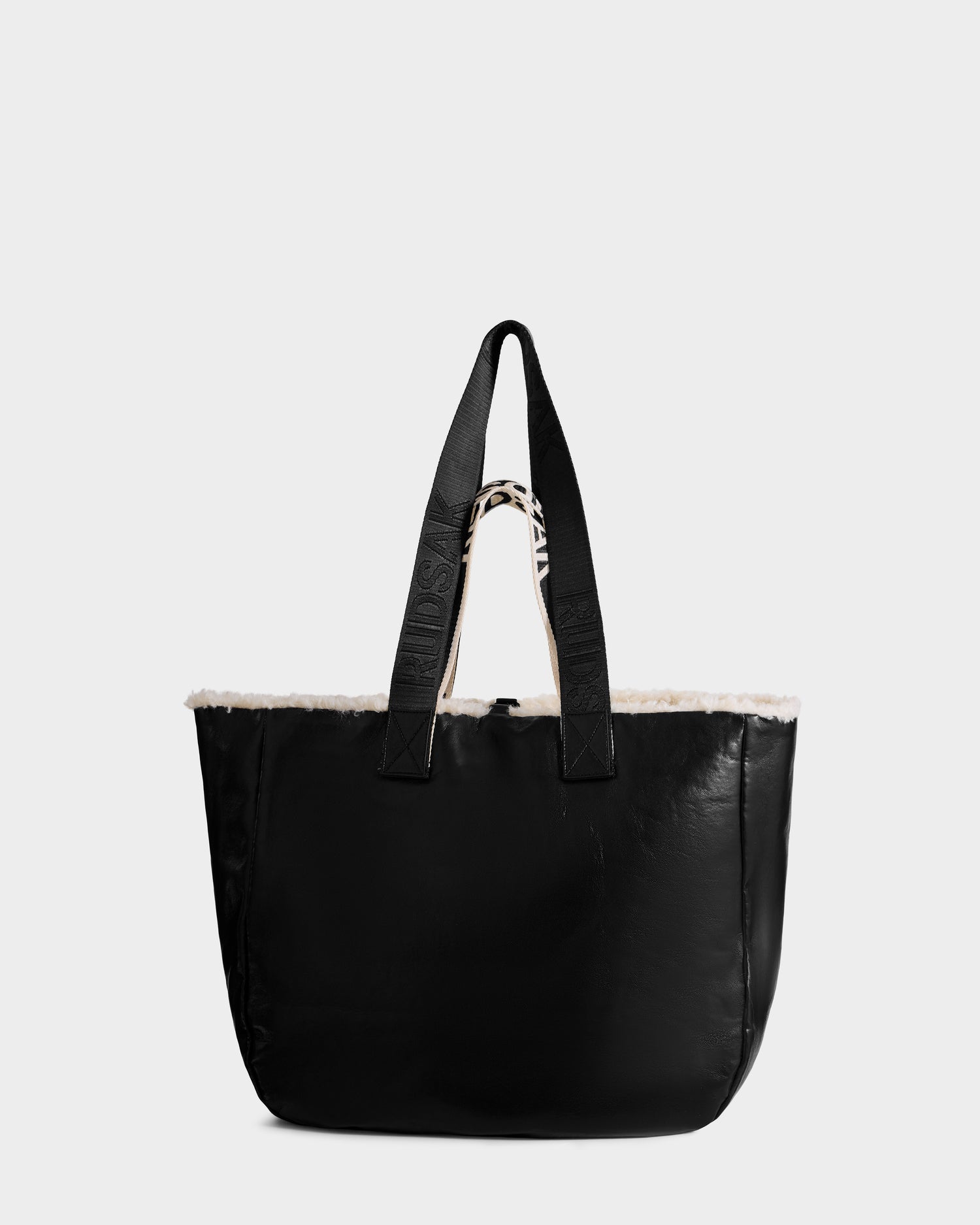 The ANNIAH - 8325541 Black/Vanilla tote bag by Rudsak features sturdy handles and a plush white interior lining, crafted in black faux shearling and photographed against a plain light gray background.