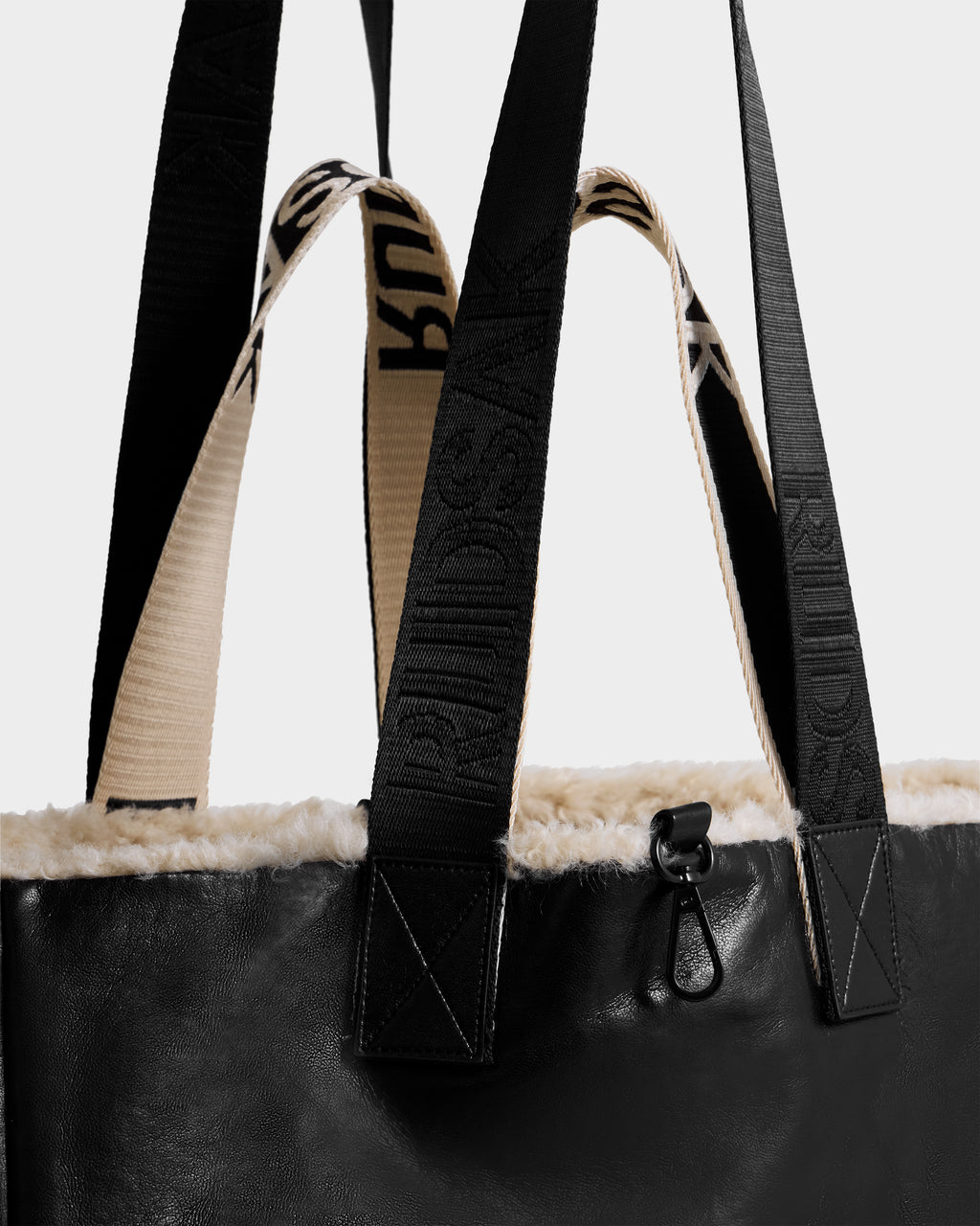 Close-up of the Rudsak ANNIAH - 8325541 tote bag in Black/Vanilla, crafted from recycled leather with cream-colored faux shearling trim and two handles—one black with embossed text, the other cream with black text. A small black metal clip accents the top edge.