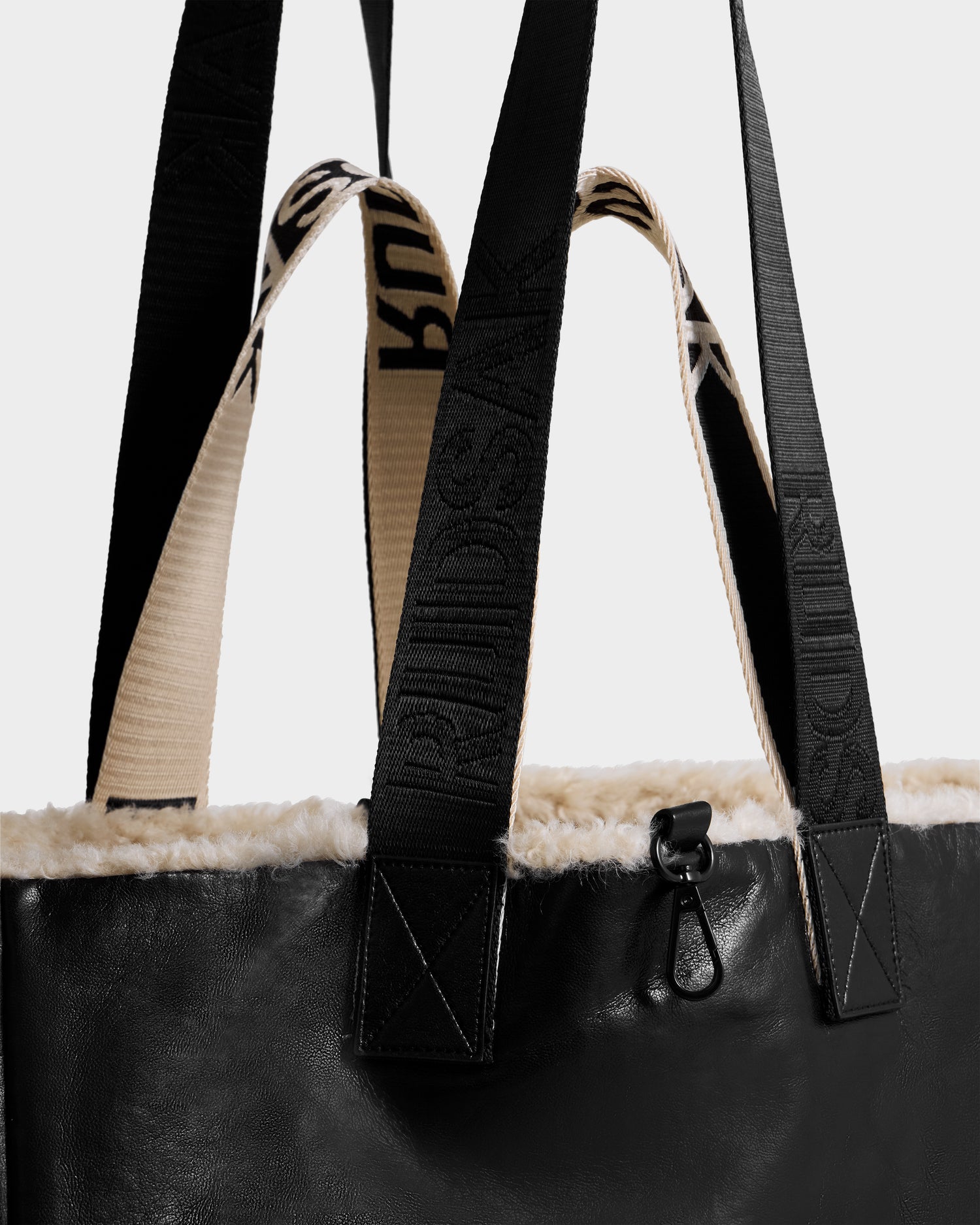 Close-up of the Rudsak ANNIAH - 8325541 tote bag in Black/Vanilla, crafted from recycled leather with cream-colored faux shearling trim and two handles—one black with embossed text, the other cream with black text. A small black metal clip accents the top edge.