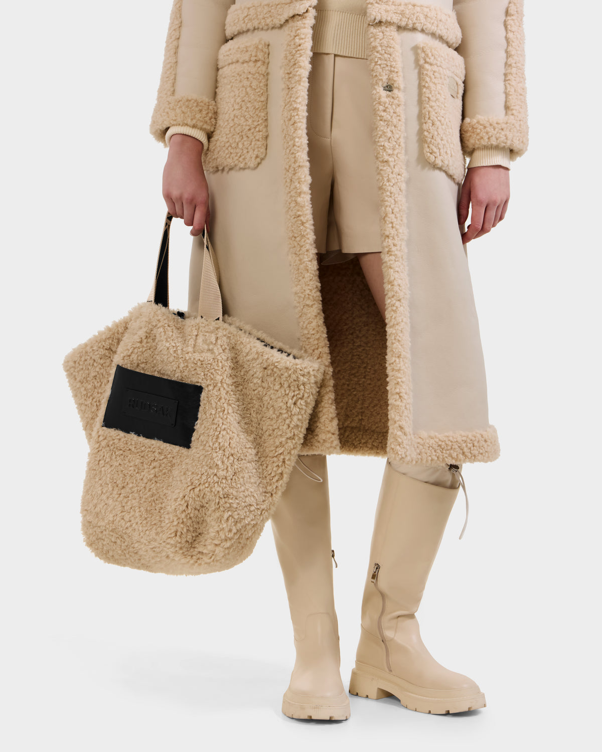 A person is shown from the waist down wearing a beige shearling coat, matching shorts, and tall beige boots, holding the Rudsak ANNIAH - 8325541 Black/Vanilla large faux shearling tote with a black patch.