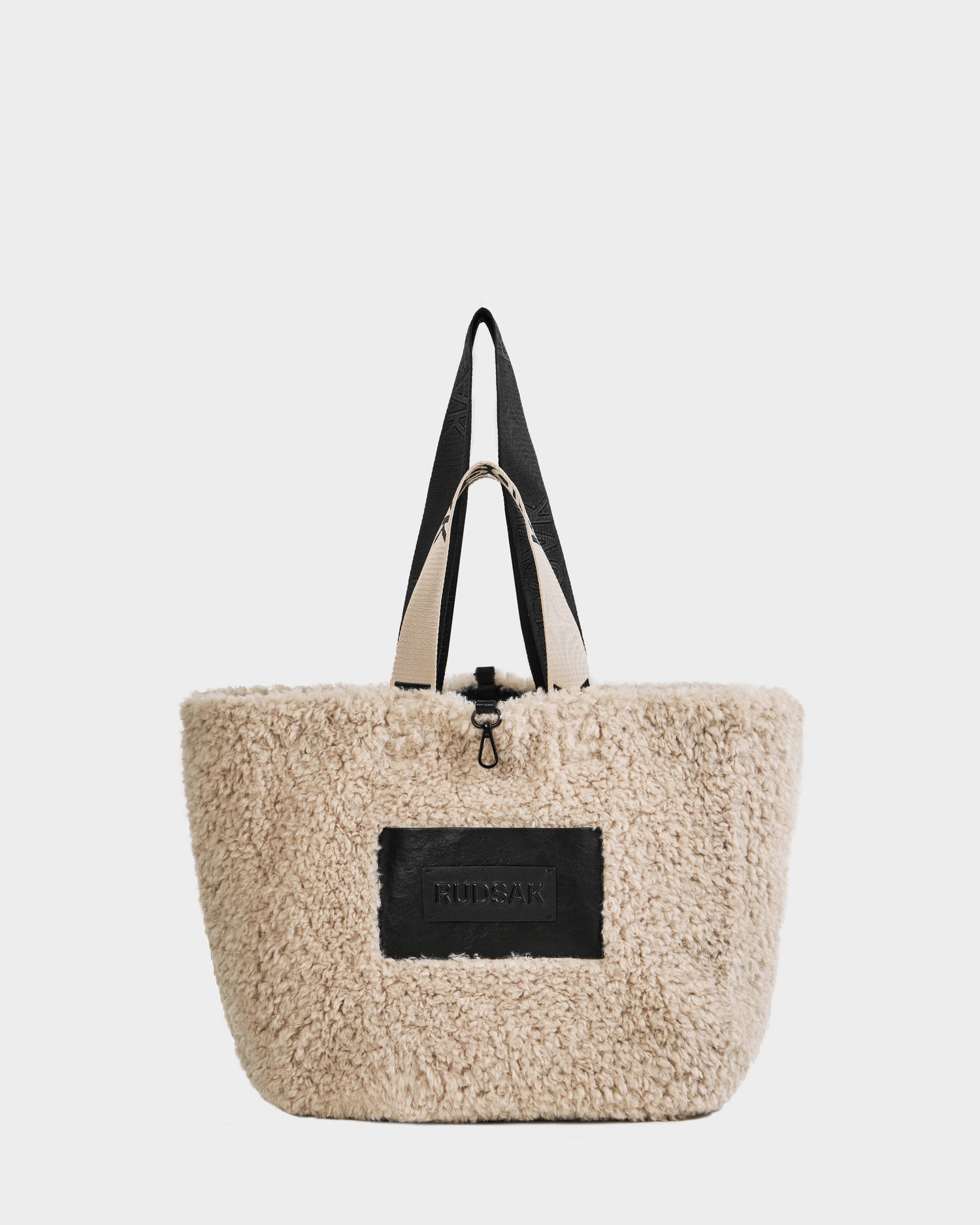 The ANNIAH - 8325541 Black/Vanilla tote bag by Rudsak features a beige faux shearling exterior with black handles and a front black patch embossed with text. This soft, fluffy sherpa tote is finished with a secure zipper closure at the top.