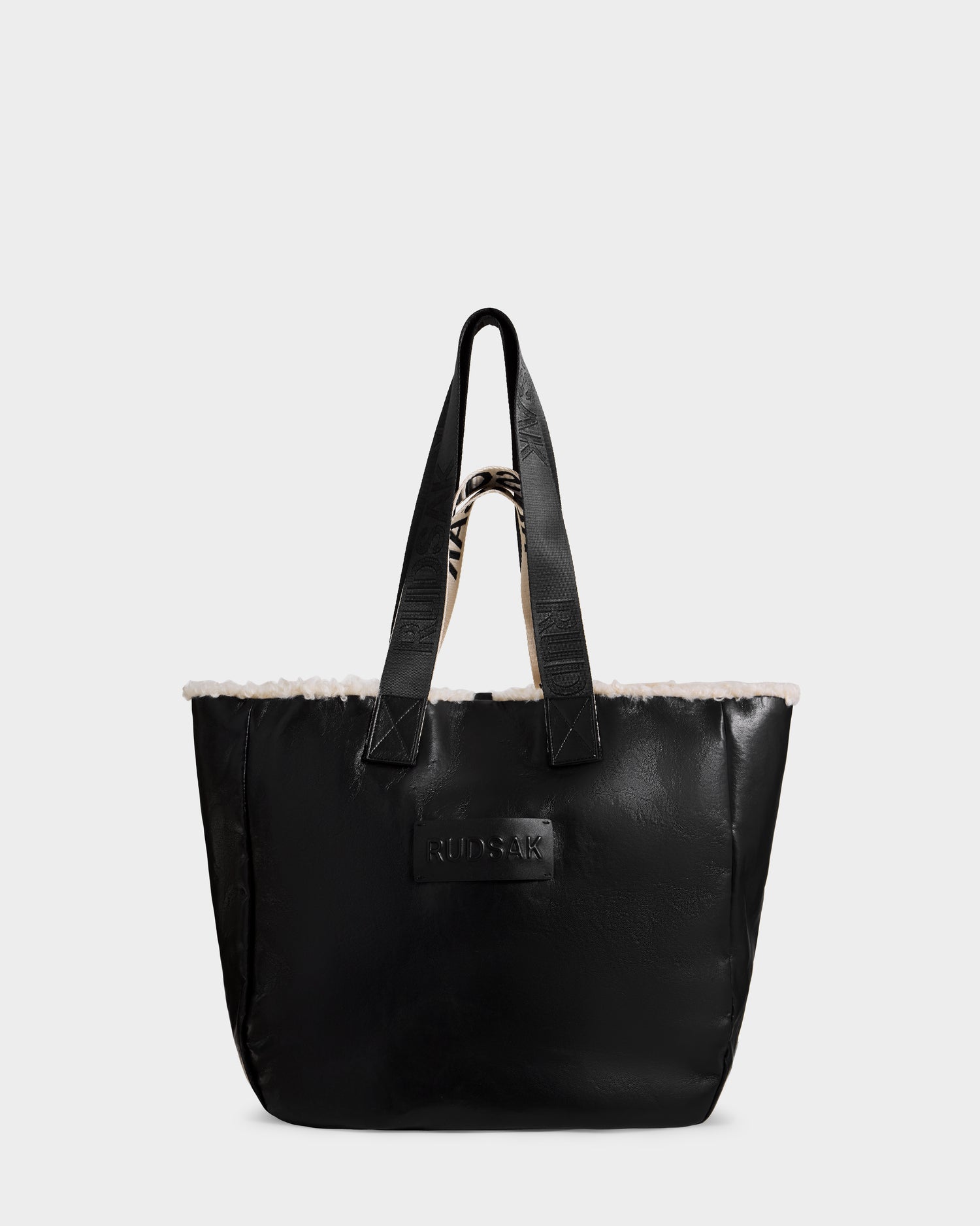 The ANNIAH - 8325541 Black/Vanilla tote bag by Rudsak features a black faux shearling design with short handles, a textured exterior, and a white fluffy trim along the top edge. A rectangular patch with embossed text decorates the front, and the bag is photographed against a plain light-colored background.