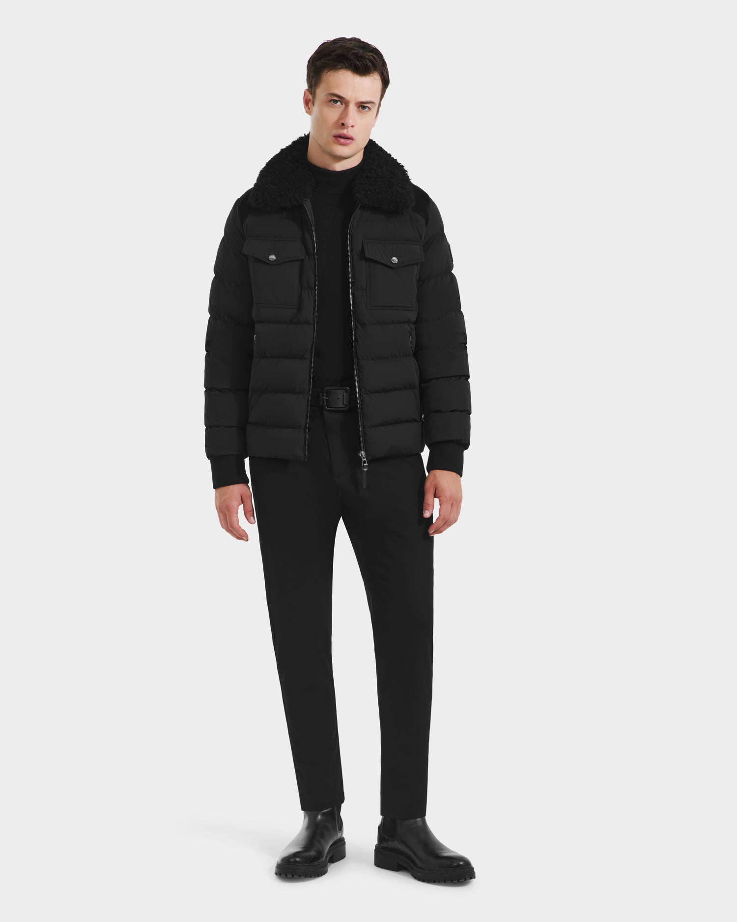 A man stands against a plain background wearing the Rudsak ANTHONY - 6125042 Black men’s aviator jacket with a shearling collar, paired with a black shirt, black pants, and black boots.
