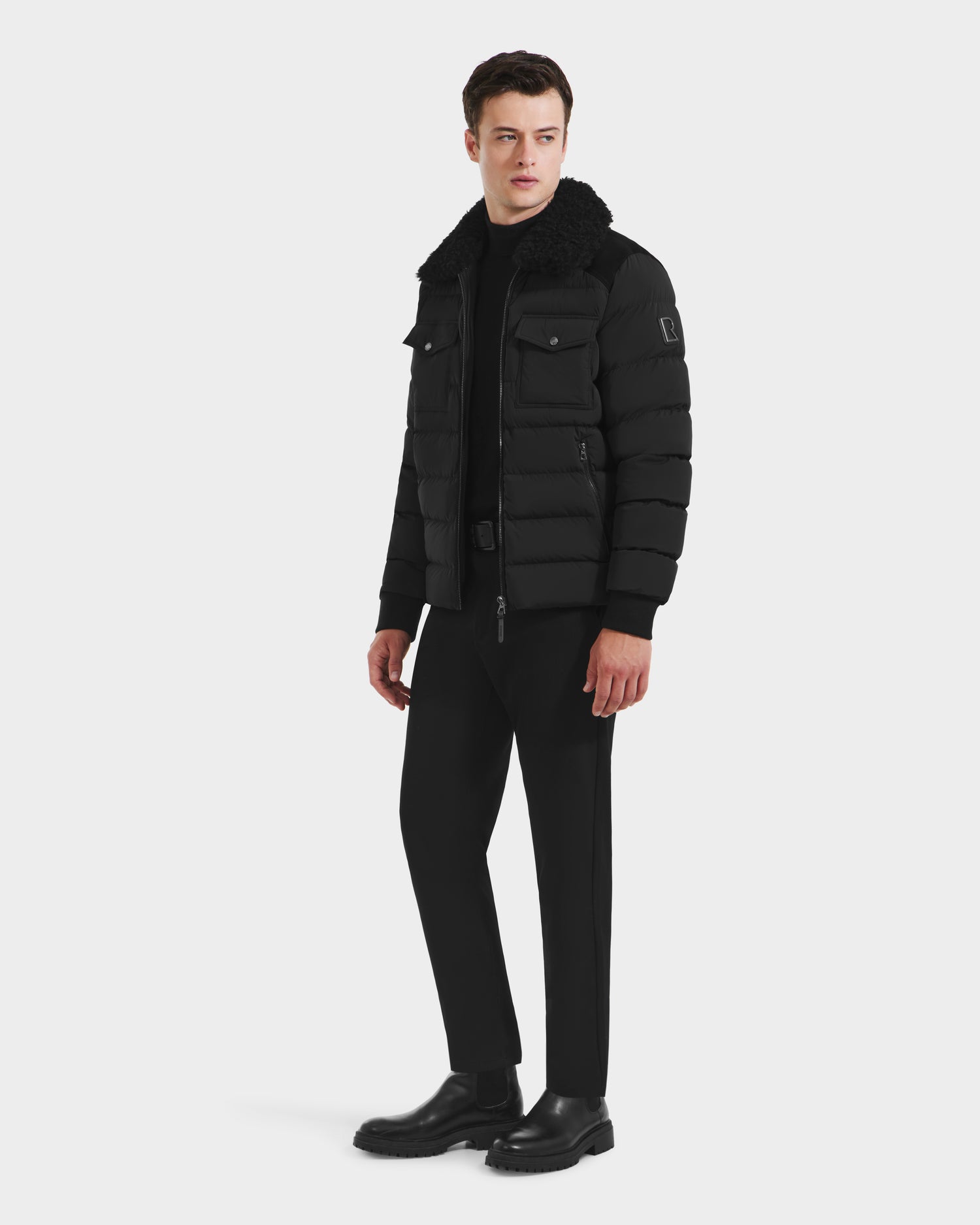A man stands against a white background wearing the Rudsak ANTHONY - 6125042 Black quilted jacket with a shearling collar, paired with black pants and boots. He faces slightly to the side with a neutral expression.