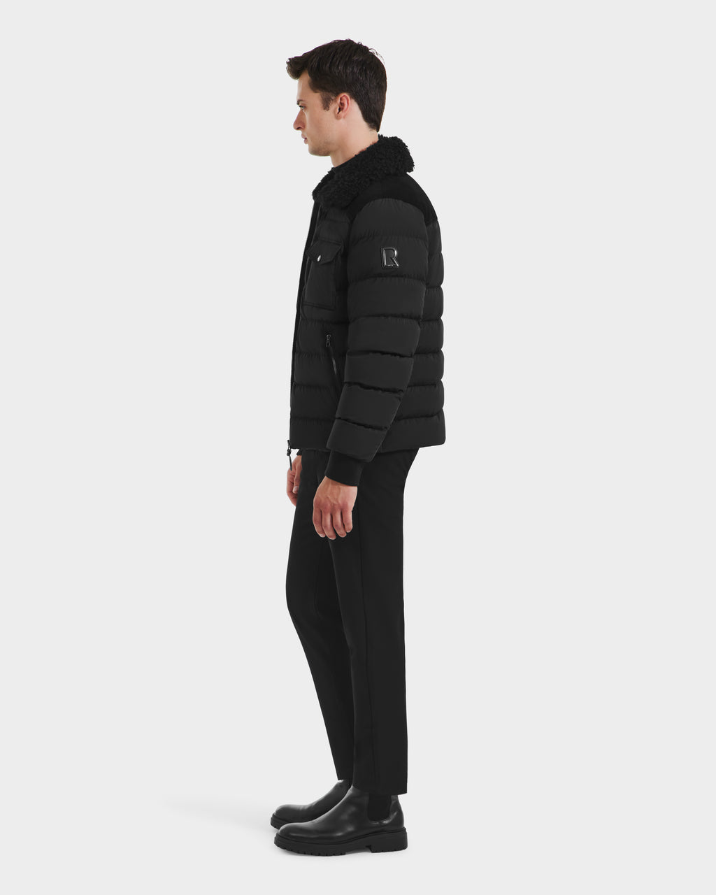 A man stands in profile against a plain white background, wearing the Rudsak ANTHONY - 6125042 Black men’s aviator jacket with a shearling collar, paired with black pants and boots.