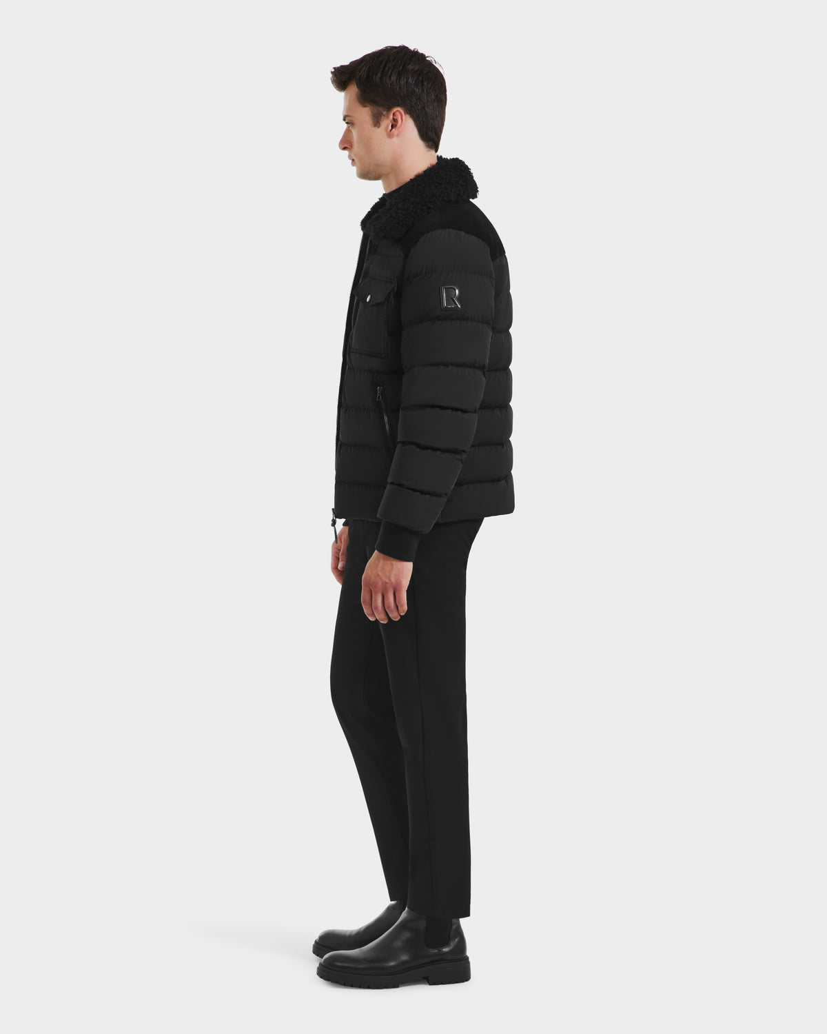 A man stands in profile against a plain white background, wearing the Rudsak ANTHONY - 6125042 Black men’s aviator jacket with a shearling collar, paired with black pants and boots.