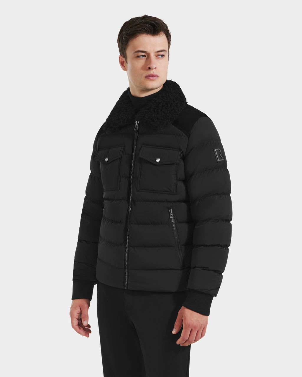 A man wearing the Rudsak ANTHONY - 6125042 Black mens aviator jacket with a shearling collar, front zip, and chest pockets stands against a plain white background, looking slightly to his left.