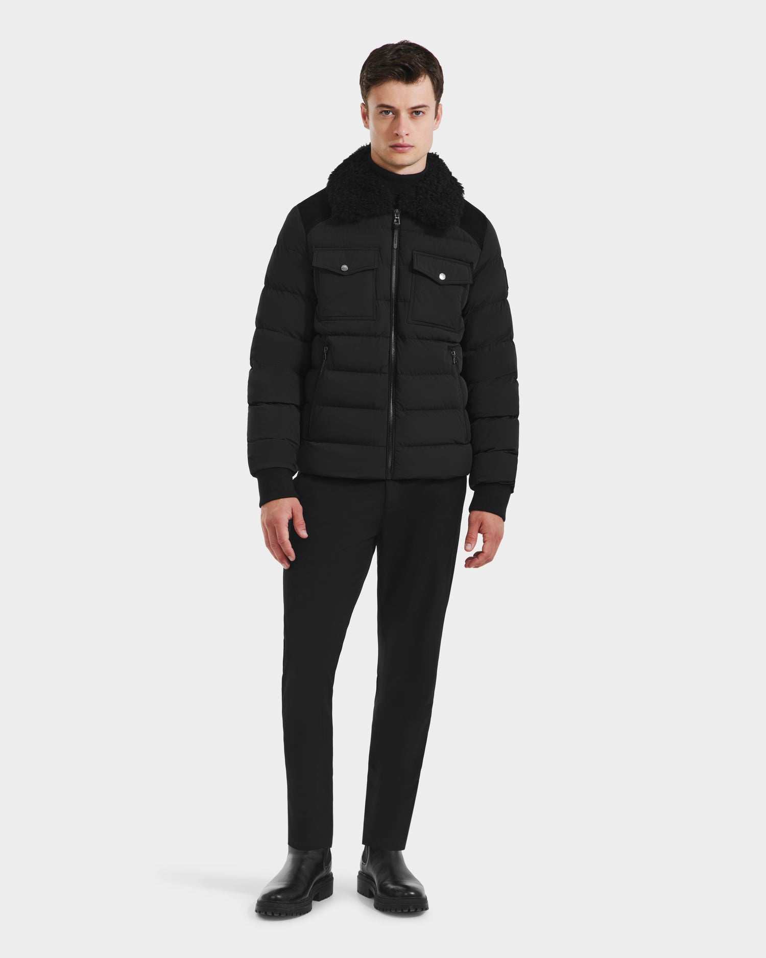 A man faces forward against a plain white background, wearing the Rudsak ANTHONY - 6125042 Black quilted jacket with a shearling collar, paired with black pants and shoes.