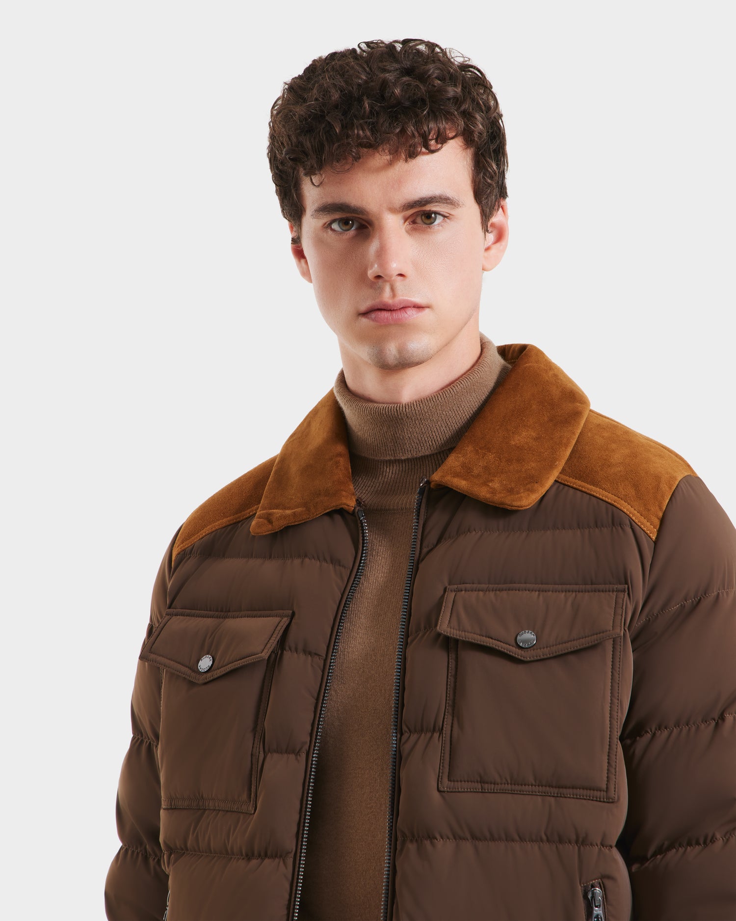 A young man with short, curly brown hair wears the Rudsak ANTHONY - 6125042 Mocha men’s aviator jacket, featuring a shearling collar and suede shoulder accents, over a tan turtleneck against a plain white background.