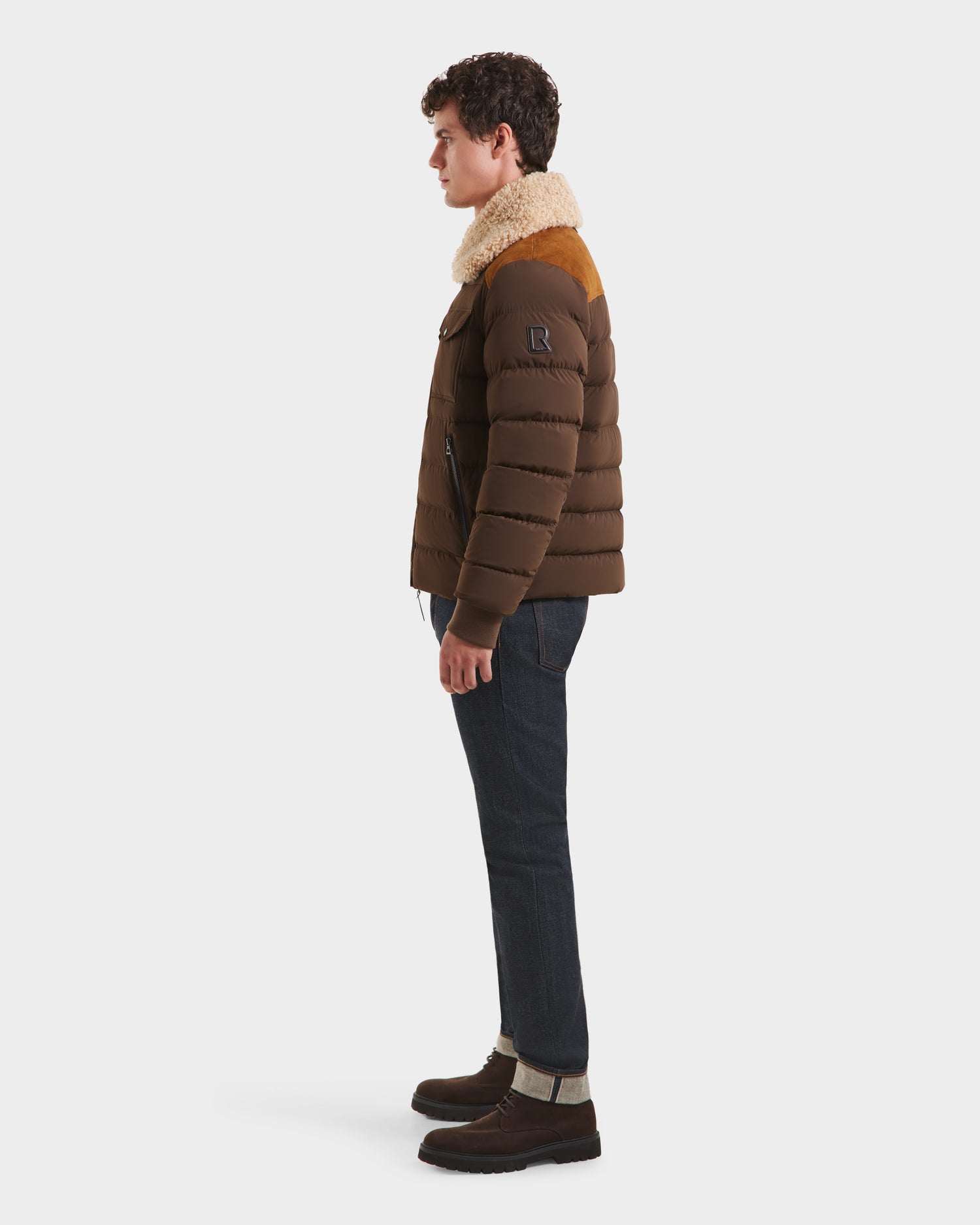 A man stands in profile against a plain background, wearing the Rudsak ANTHONY - 6125042 Mocha aviator jacket with a shearling collar, paired with dark jeans and brown boots.