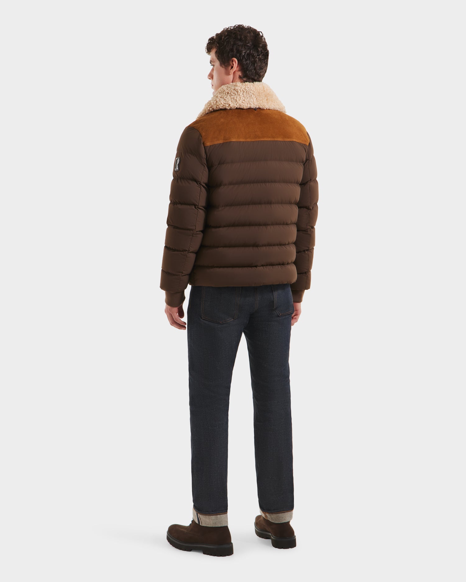A man faces away wearing the Rudsak ANTHONY - 6125042 Mocha men’s aviator jacket with a shearling collar, dark jeans, and brown shoes, set against a plain white background.
