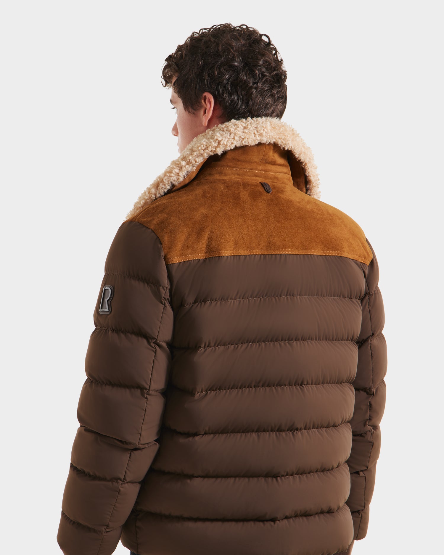 A person with curly hair is seen from behind wearing the Rudsak ANTHONY - 6125042 Mocha jacket, featuring a brown quilted design, tan suede yoke, shearling collar, and an R patch on the upper left sleeve.