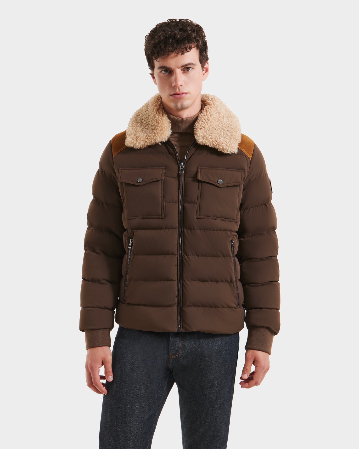 A young man wears the Rudsak ANTHONY - 6125042 Mocha quilted jacket with a beige shearling collar and chest pockets, paired with dark jeans, standing against a plain white background.