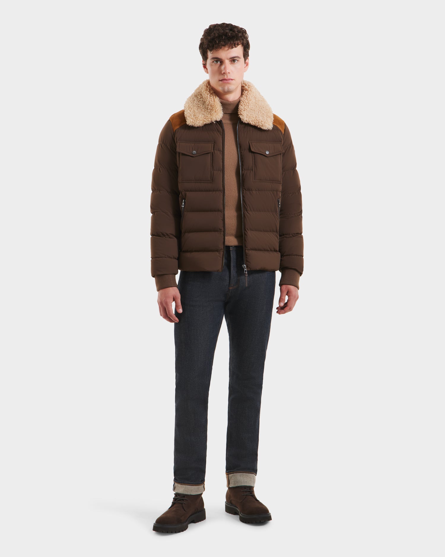 A man faces forward against a plain white background, wearing the Rudsak ANTHONY - 6125042 Mocha quilted jacket with a tan shearling collar, a tan sweater, dark rolled-cuff jeans, and brown shoes.