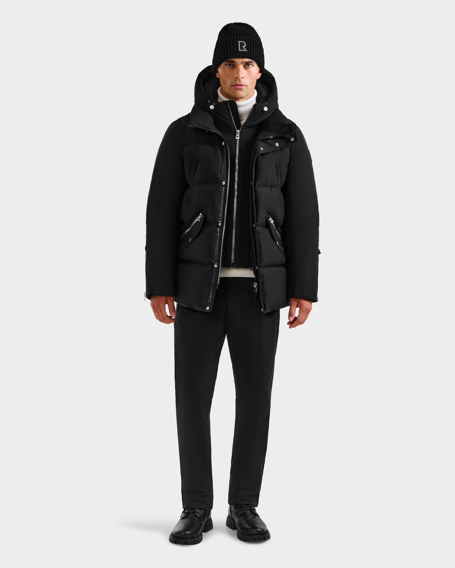 A person faces forward against a light gray background, wearing a Rudsak APOLLO H - 6125690 Black men's down parka, paired with black pants, black shoes, and a white turtleneck sweater.
