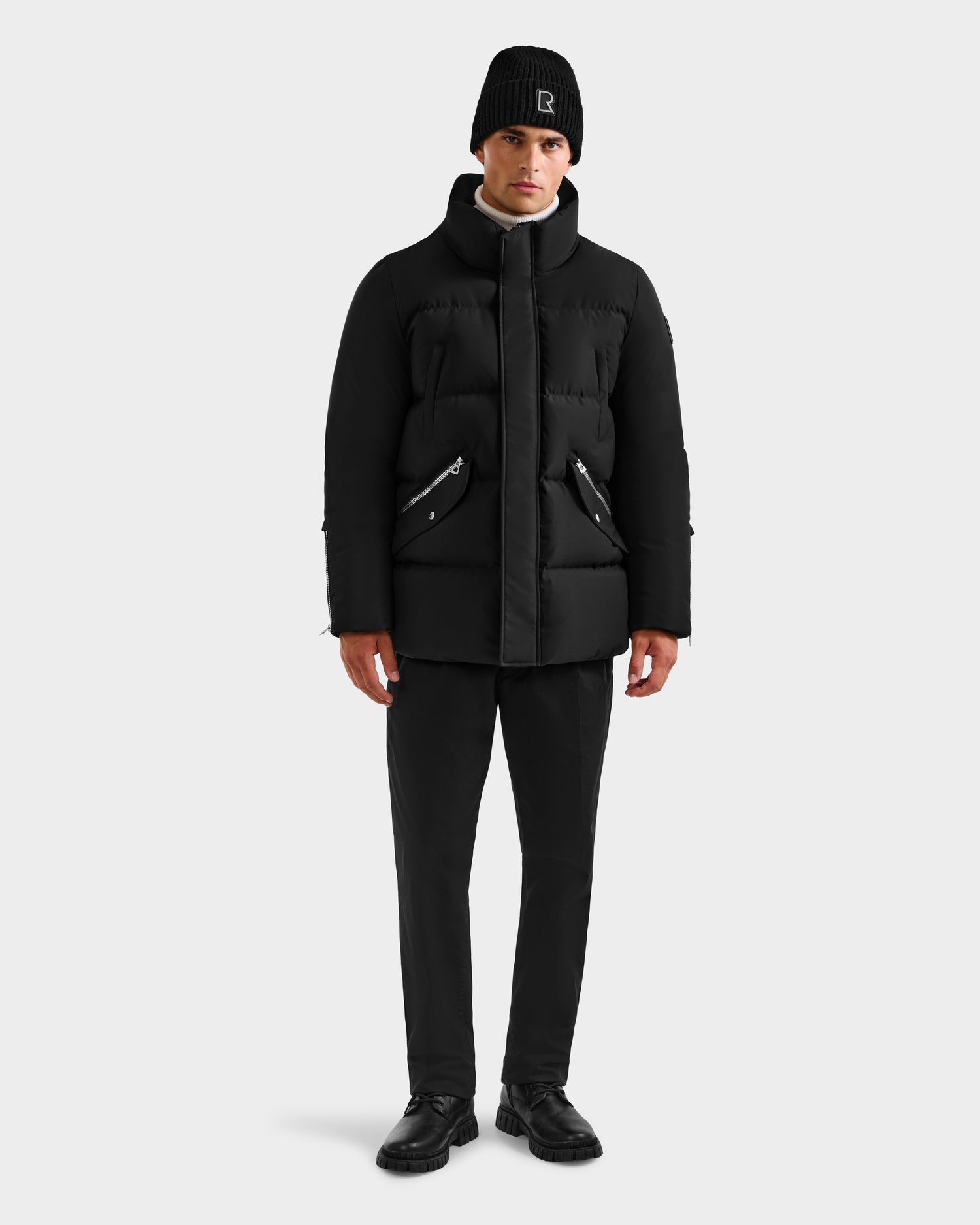 A man stands facing forward wearing the Rudsak APOLLO H - 6125690 Black men’s quilted down parka, paired with black pants and shoes, against a plain light background.