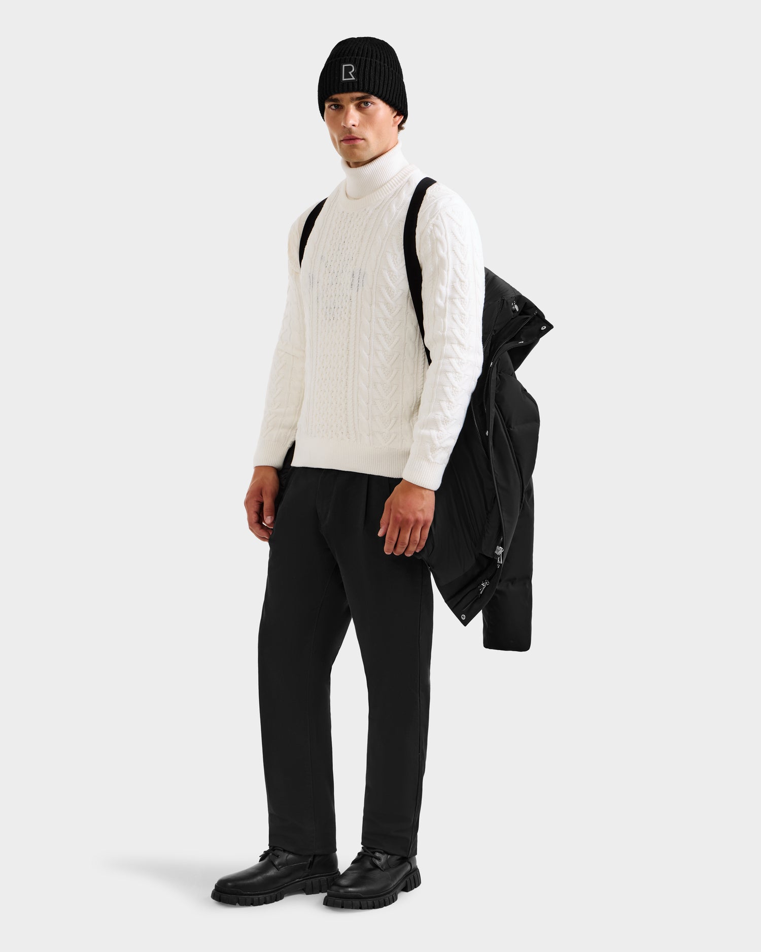A man in a white cable-knit turtleneck, black pants, winter hat, and boots stands holding the Rudsak APOLLO H - 6125690 Black men’s down parka over his shoulder against a plain light background, wearing a neutral expression.
