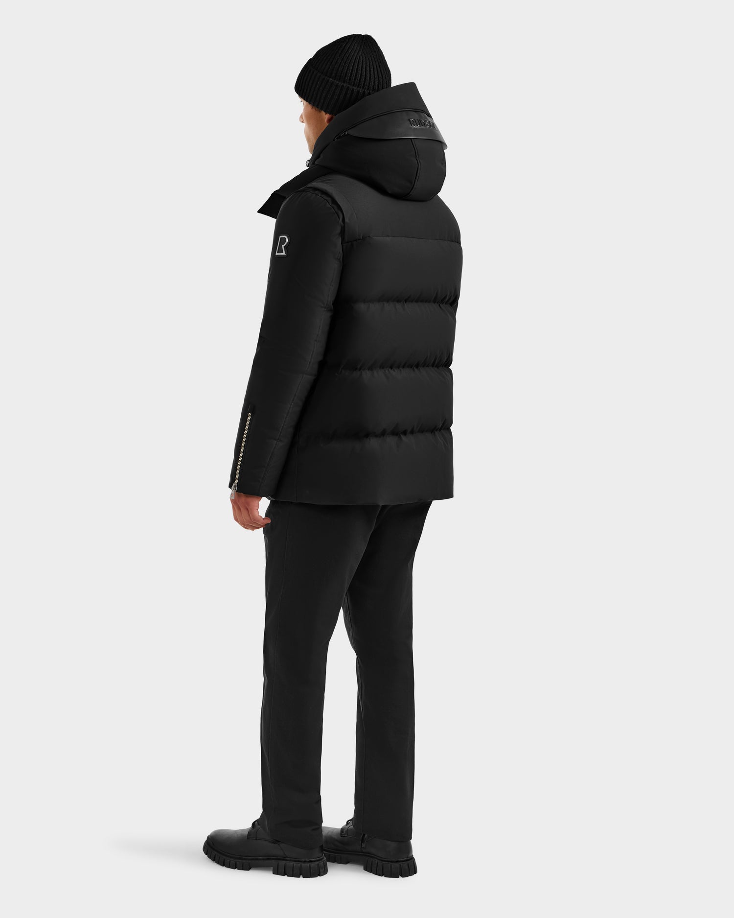 A person wearing the Rudsak APOLLO H - 6125690 Black quilted down parka, black beanie, black pants, and black boots stands facing away against a plain light background.