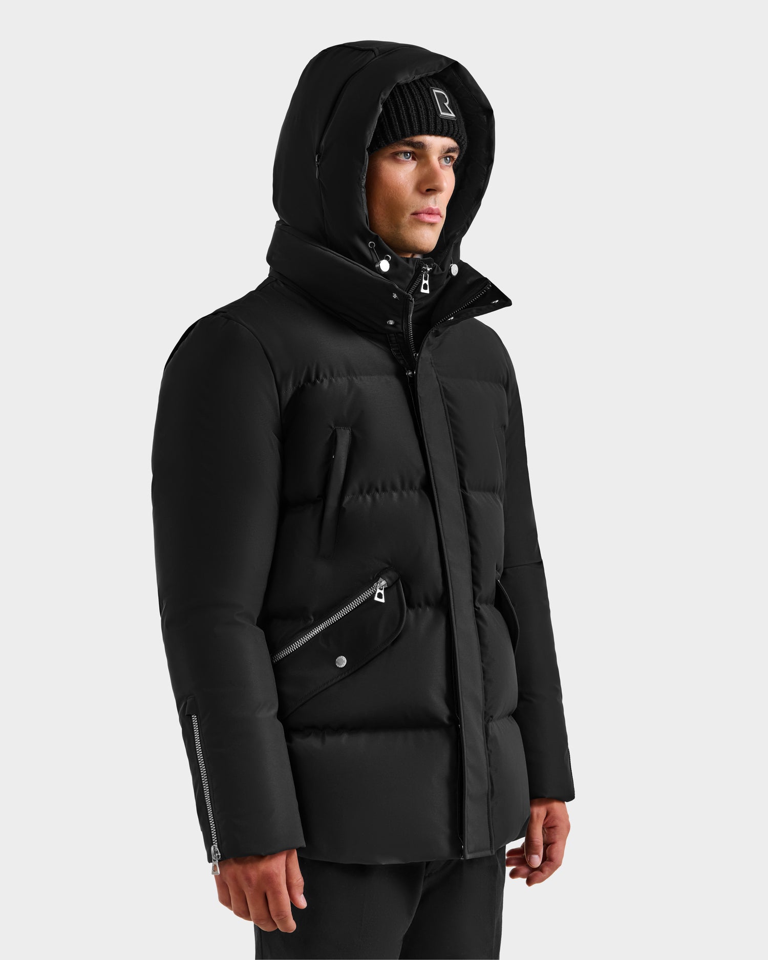 A man in the Rudsak APOLLO H - 6125690 Black quilted down parka with hood and zipper details, paired with a black beanie, stands slightly turned to the side against a plain light background.