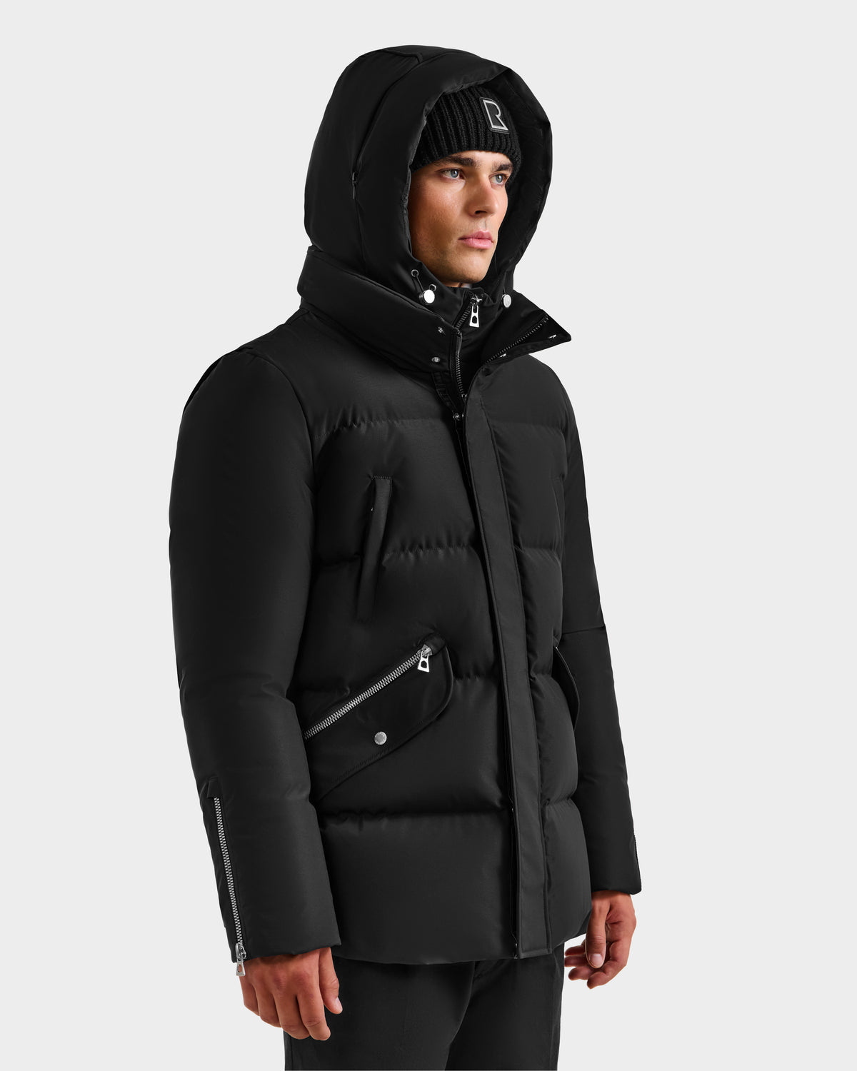 A man in the Rudsak APOLLO H - 6125690 Black quilted down parka with hood and zipper details, paired with a black beanie, stands slightly turned to the side against a plain light background.