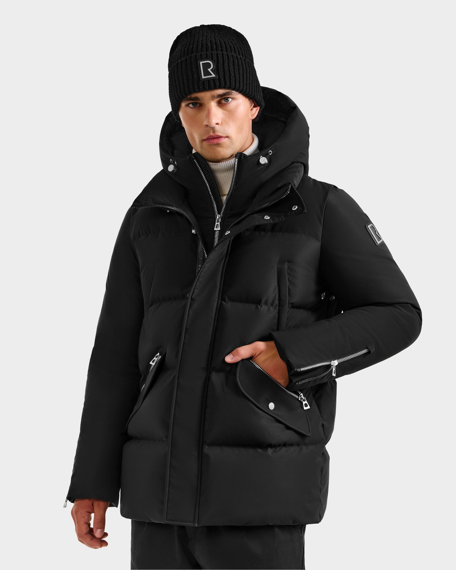 A person wears the Rudsak APOLLO H - 6125690 Black jacket with vegan leather trim and multiple zippers, paired with a black knit beanie. They face forward, left hand in pocket, against a plain light gray background.