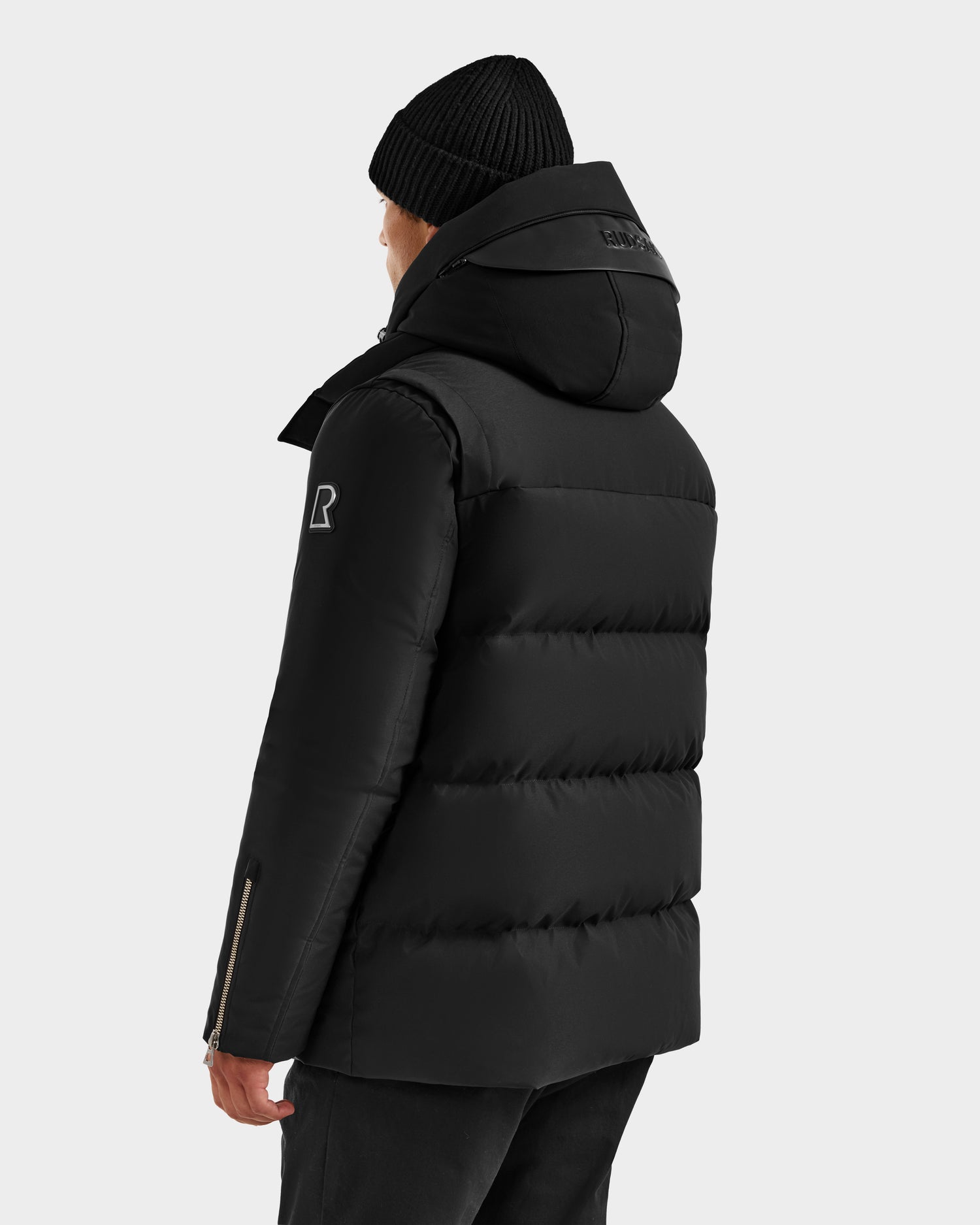 A person wearing the Rudsak APOLLO H - 6125690 Black men's down parka with a hood and a black knit beanie, seen from the back against a light background. The quilted jacket displays a visible logo on the left sleeve.