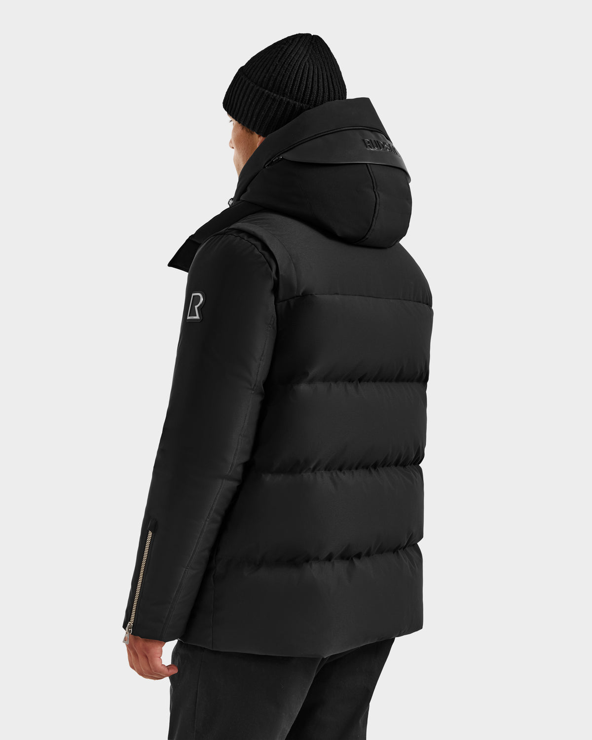 A person wearing the Rudsak APOLLO H - 6125690 Black men's down parka with a hood and a black knit beanie, seen from the back against a light background. The quilted jacket displays a visible logo on the left sleeve.