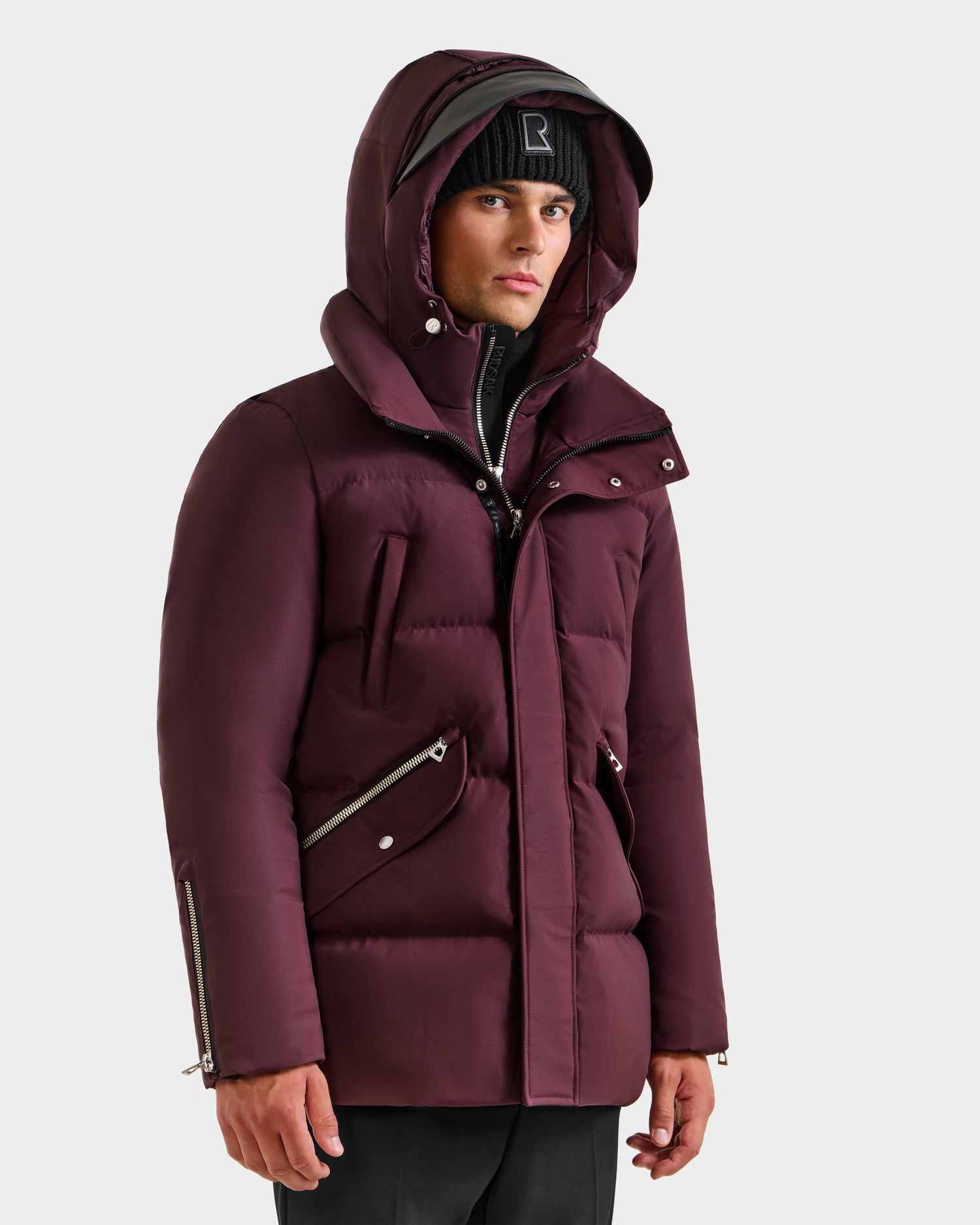 A person stands slightly to the side in a Rudsak APOLLO H - 6125690 Bordeaux parka with a hood and silver zippers, styled with a black beanie and black pants, against a plain light background.